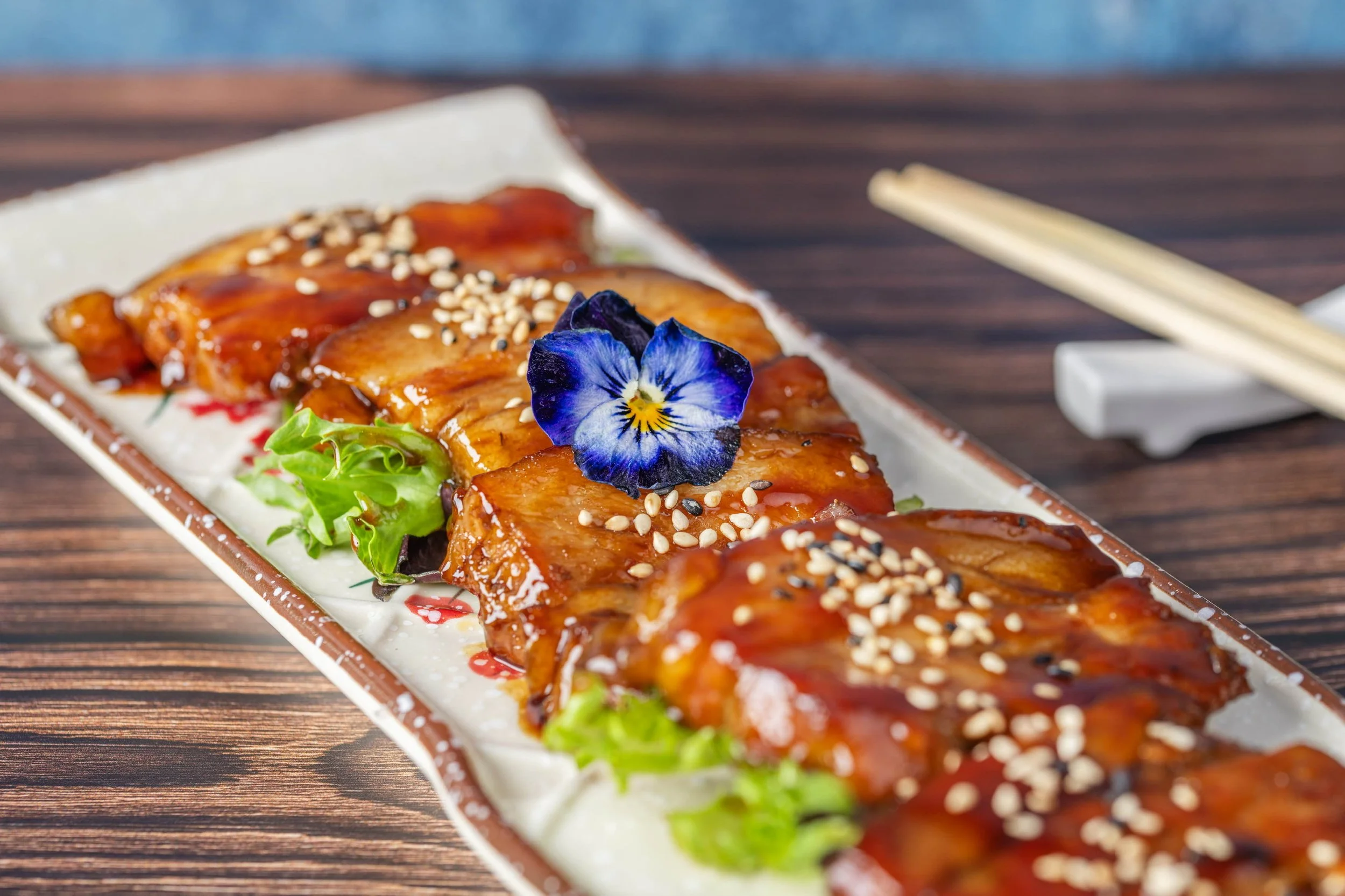Glazed Japanese dish topped with sesame seeds and an edible flower on a rectangular platter.