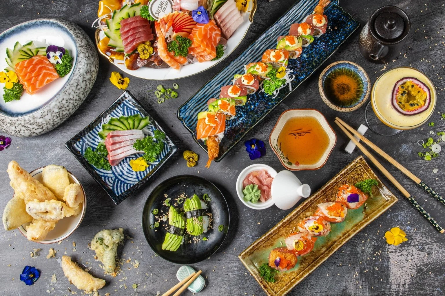 Overhead view of sushi, sashimi, tempura, vegetable dishes and drinks arranged for sharing.