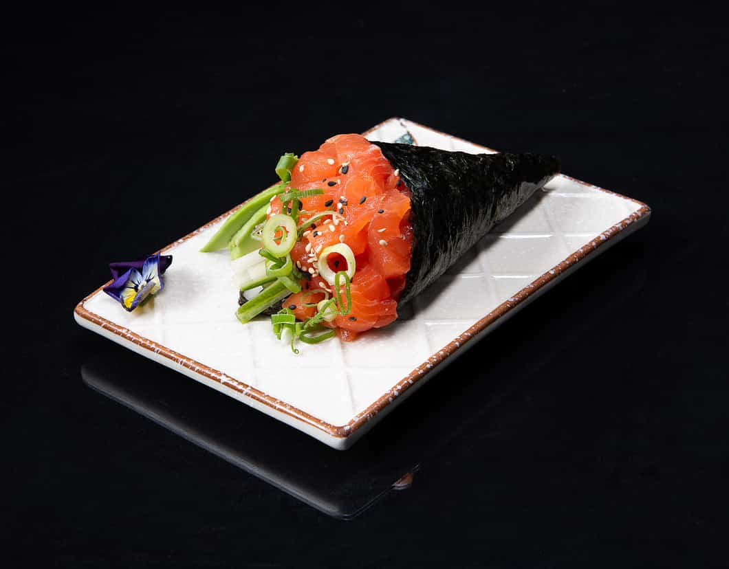 Sushi hand roll with salmon, cucumber, green onions, and sesame seeds, served on a rectangular white plate with a small flower garnish.
