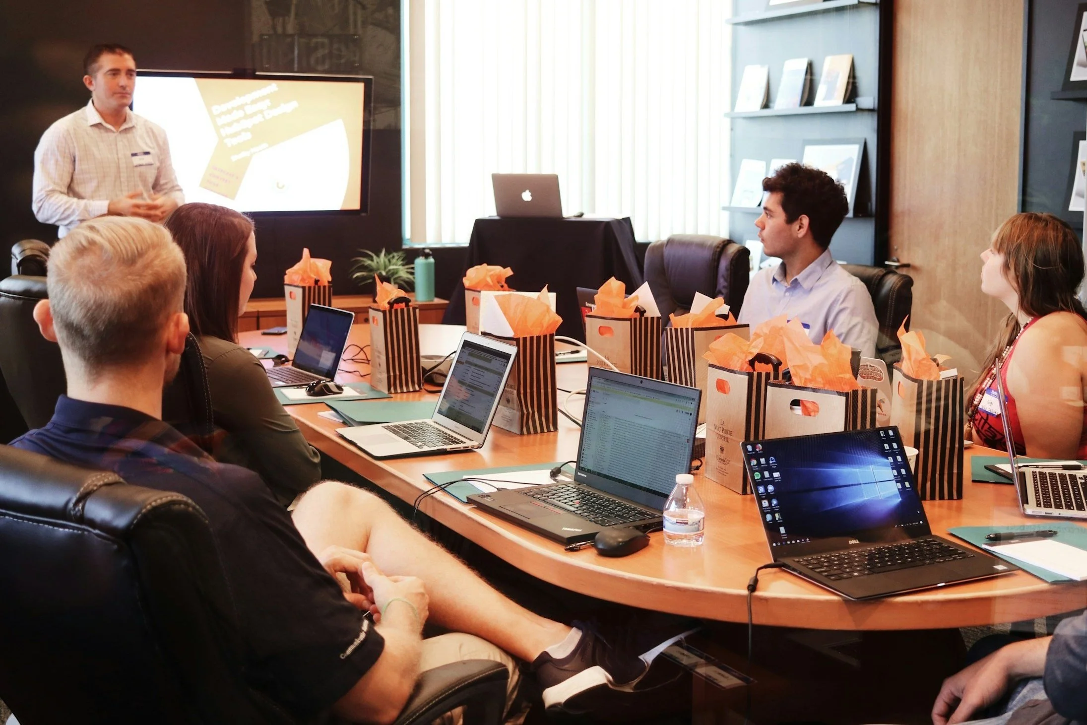 A group of people in a meeting room with a presenter at the front, presentation screen behind him, laptops, gift bags, and a water bottle on the table.