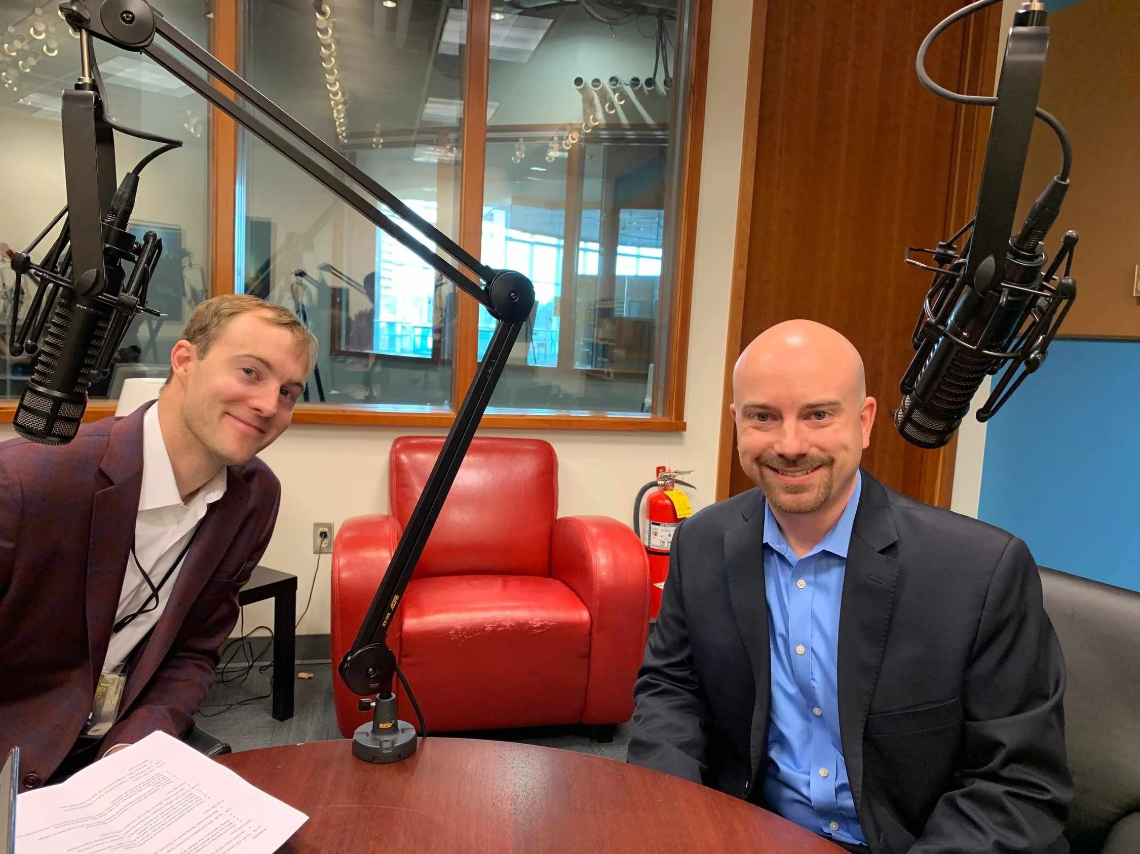 Two men are sitting at a round table in a recording studio with microphones mounted overhead. The man on the left has short light brown hair and is wearing a plaid shirt and blazer, smiling slightly. The man on the right is bald, has a beard, and is wearing a blue shirt with a dark blazer, smiling broadly. There are papers on the table and a red armchair and a fire extinguisher in the background.