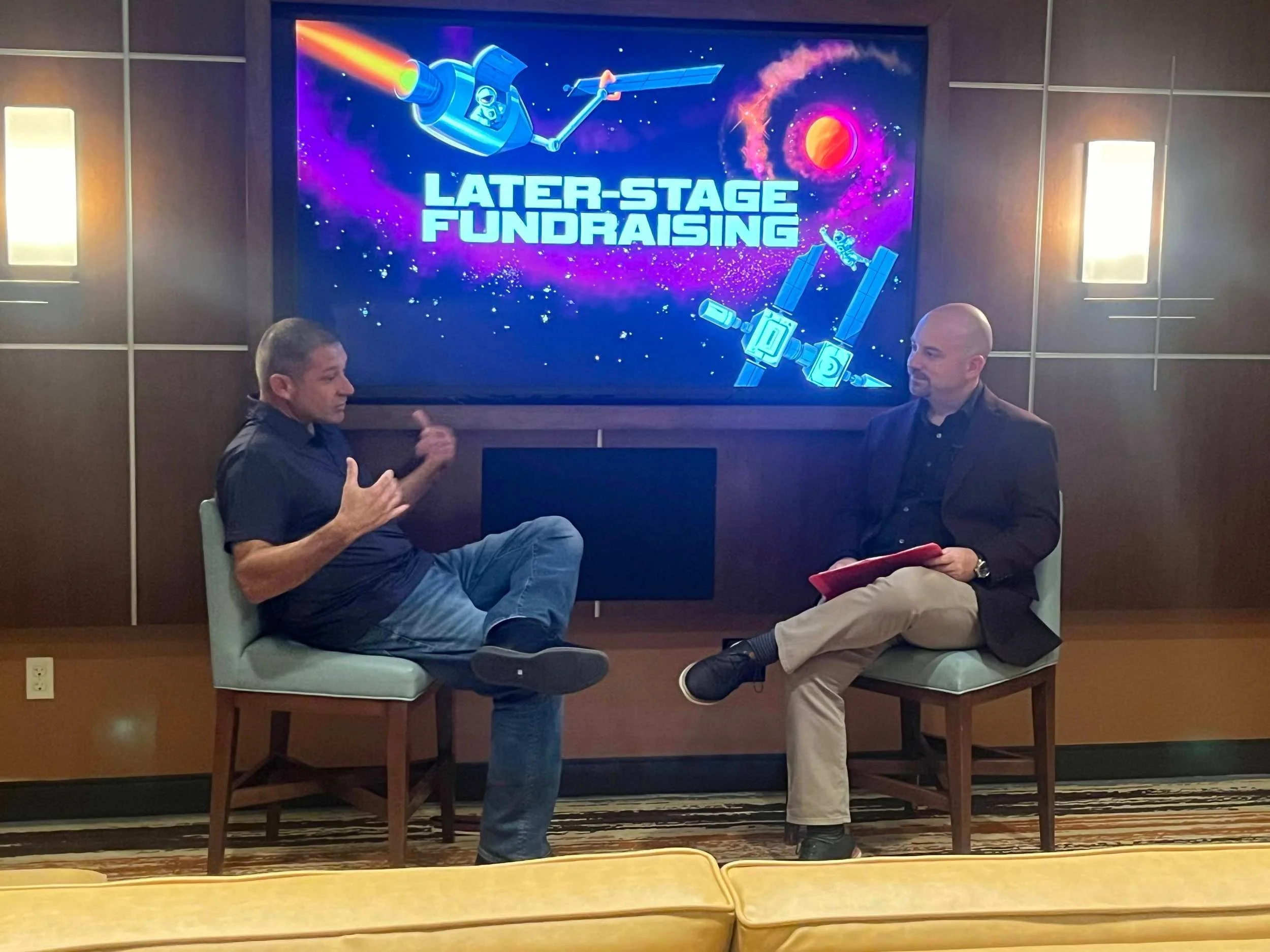 Two men are seated and talking in front of a large screen that reads 'Later-Stage Fundraising' with illustrations of a rocket and satellites in space.