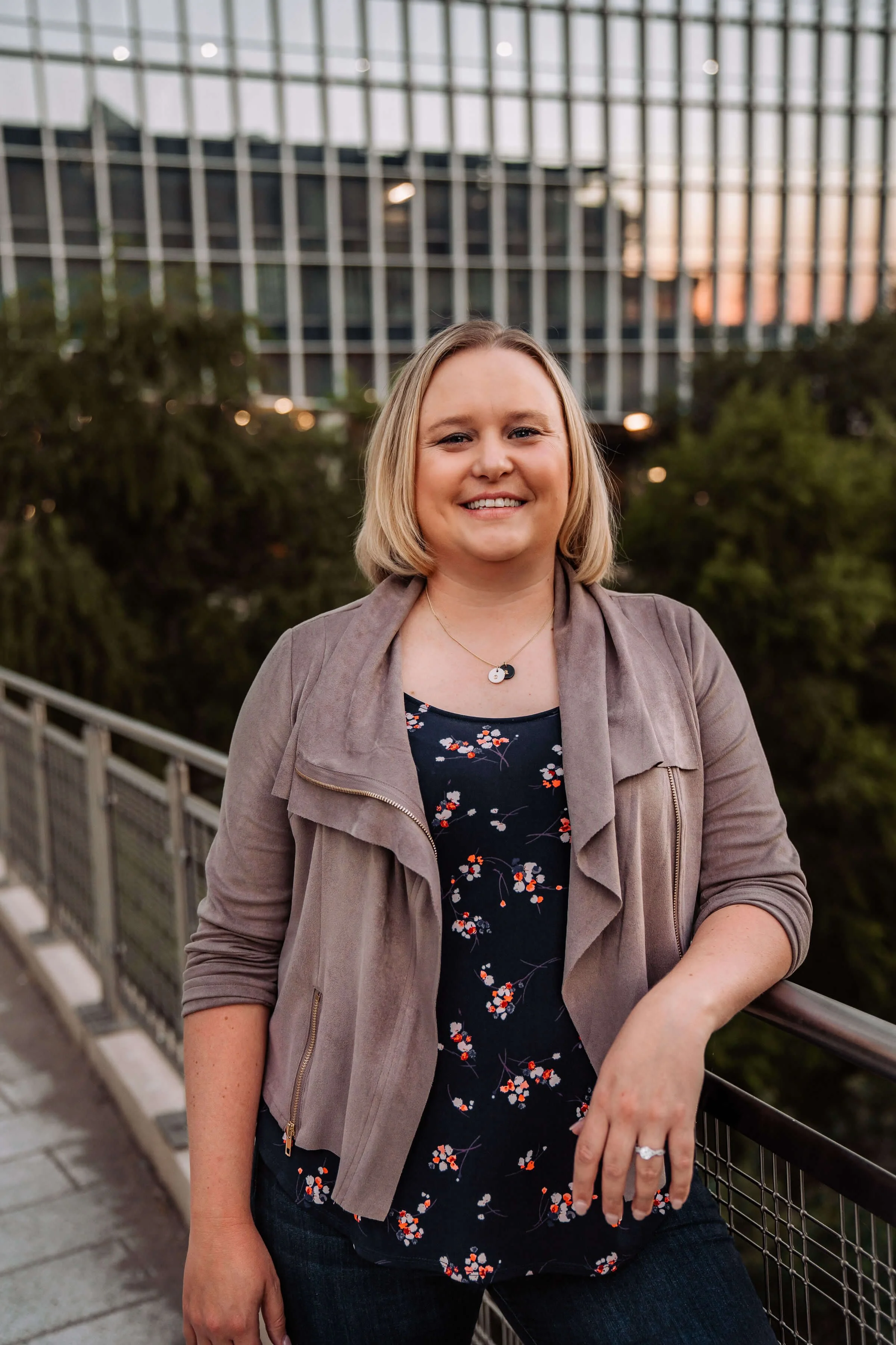 A woman with blonde hair smiling outdoors at sunset, wearing a beige jacket over a dark floral blouse, standing by a railing with a modern building in the background.