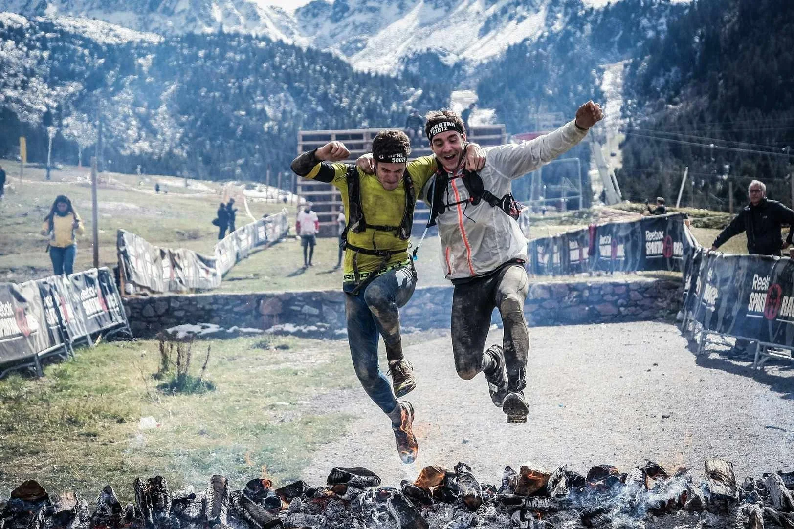 Two male runners in athletic gear and backpacks cross a finish line at a mountain race, celebrating with arms raised in a scenic outdoor setting with snow-covered mountains in the background.