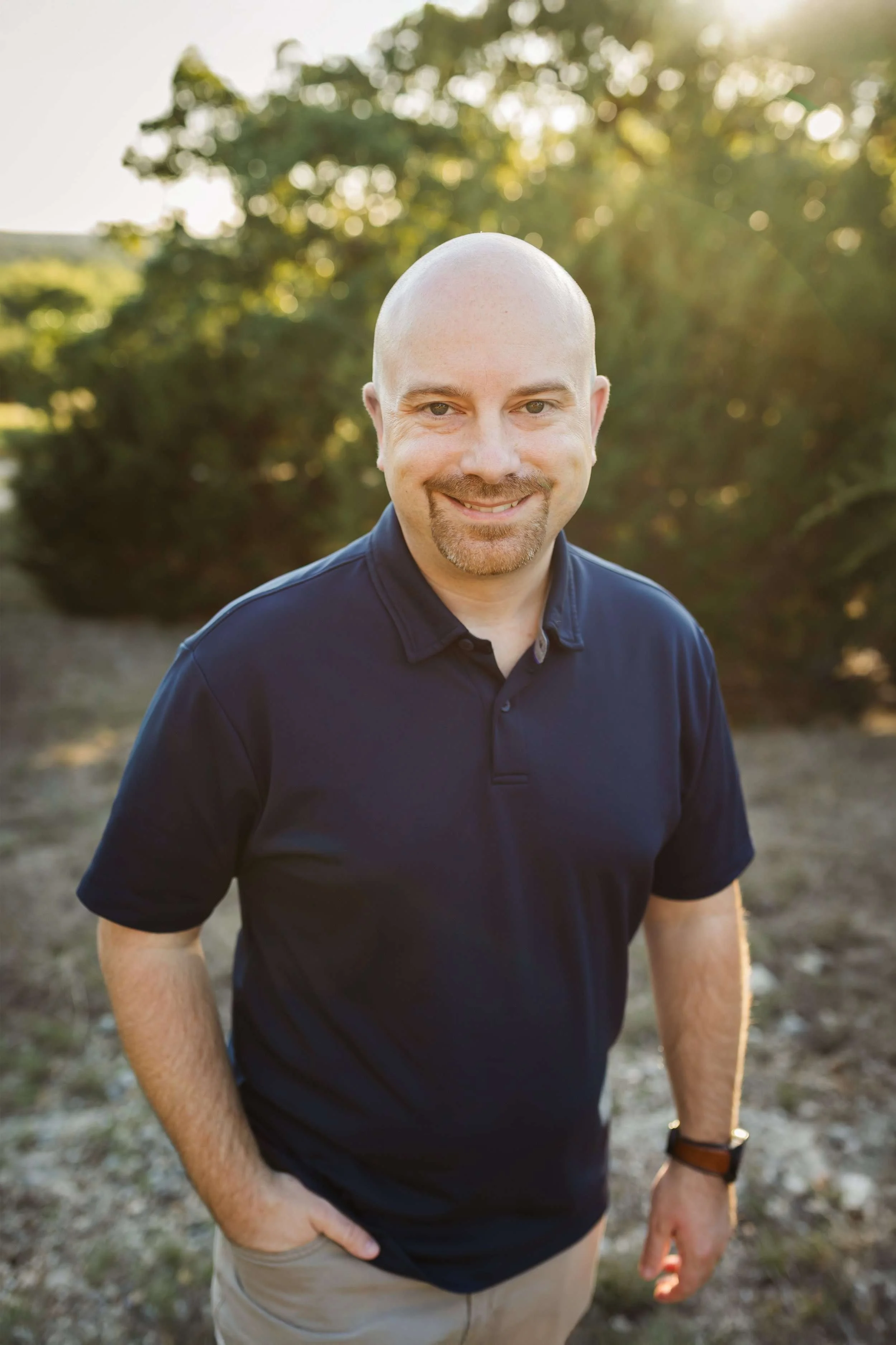A man with a beard and bald head smiling outdoors in sunlight, wearing a navy polo shirt and beige pants.