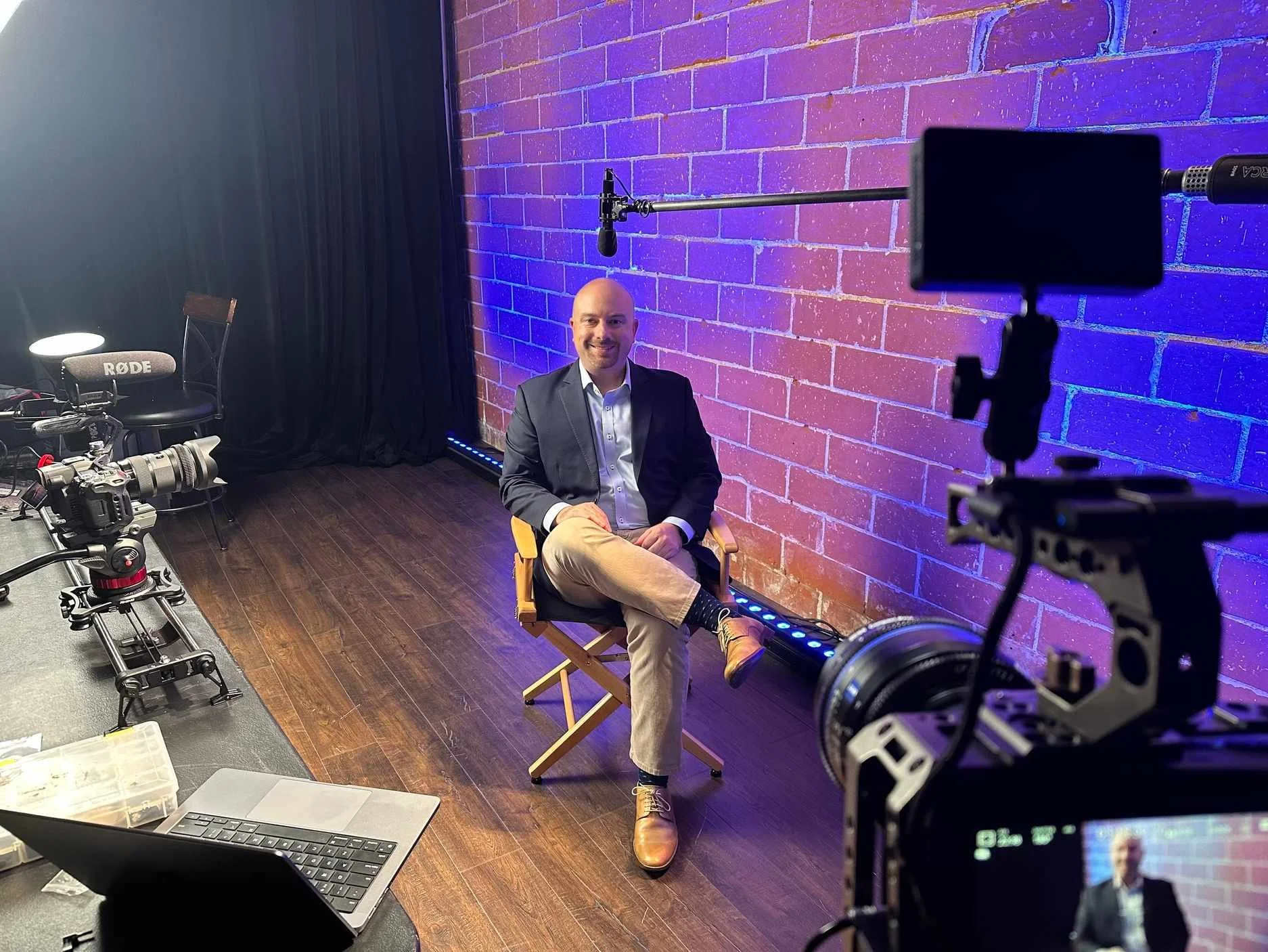A man in business attire sits on a director's chair in a recording studio with a brick wall background and colorful lighting, surrounded by professional filming equipment.
