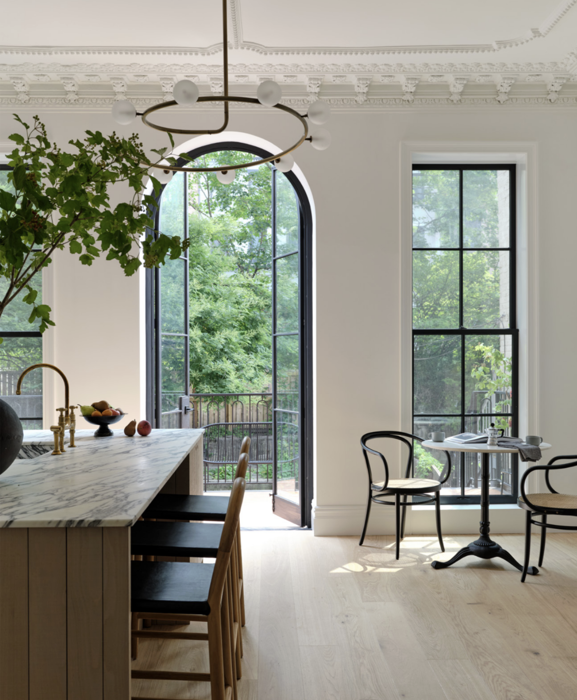 Bright kitchen with large arched window and black-framed smaller window, white walls, ornate crown molding, modern chandelier, marble kitchen island with brown chairs, small round table with two black chairs, greenery visible outside.
