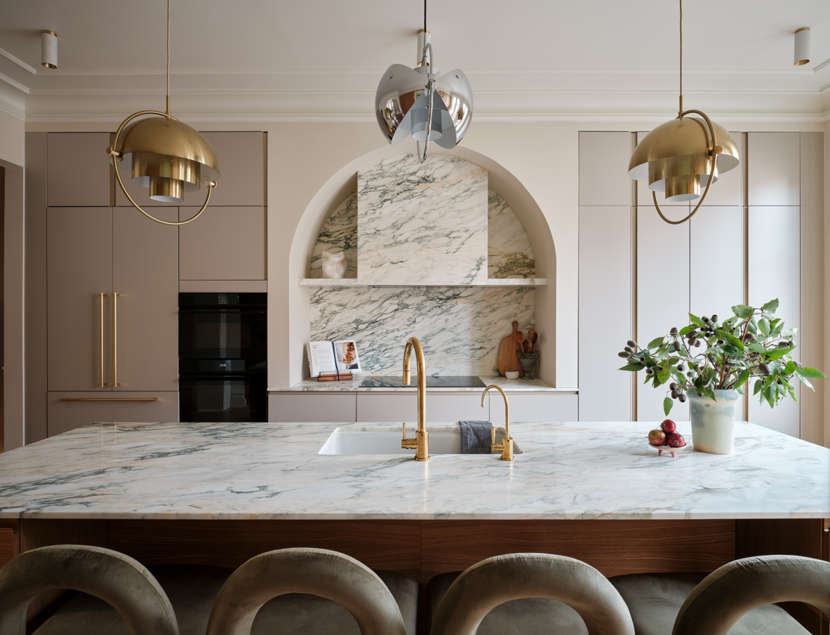 Modern kitchen with a marble island, gold faucets, beige cabinets, and unique hanging lights, with a vase of greenery and apples on the counter.