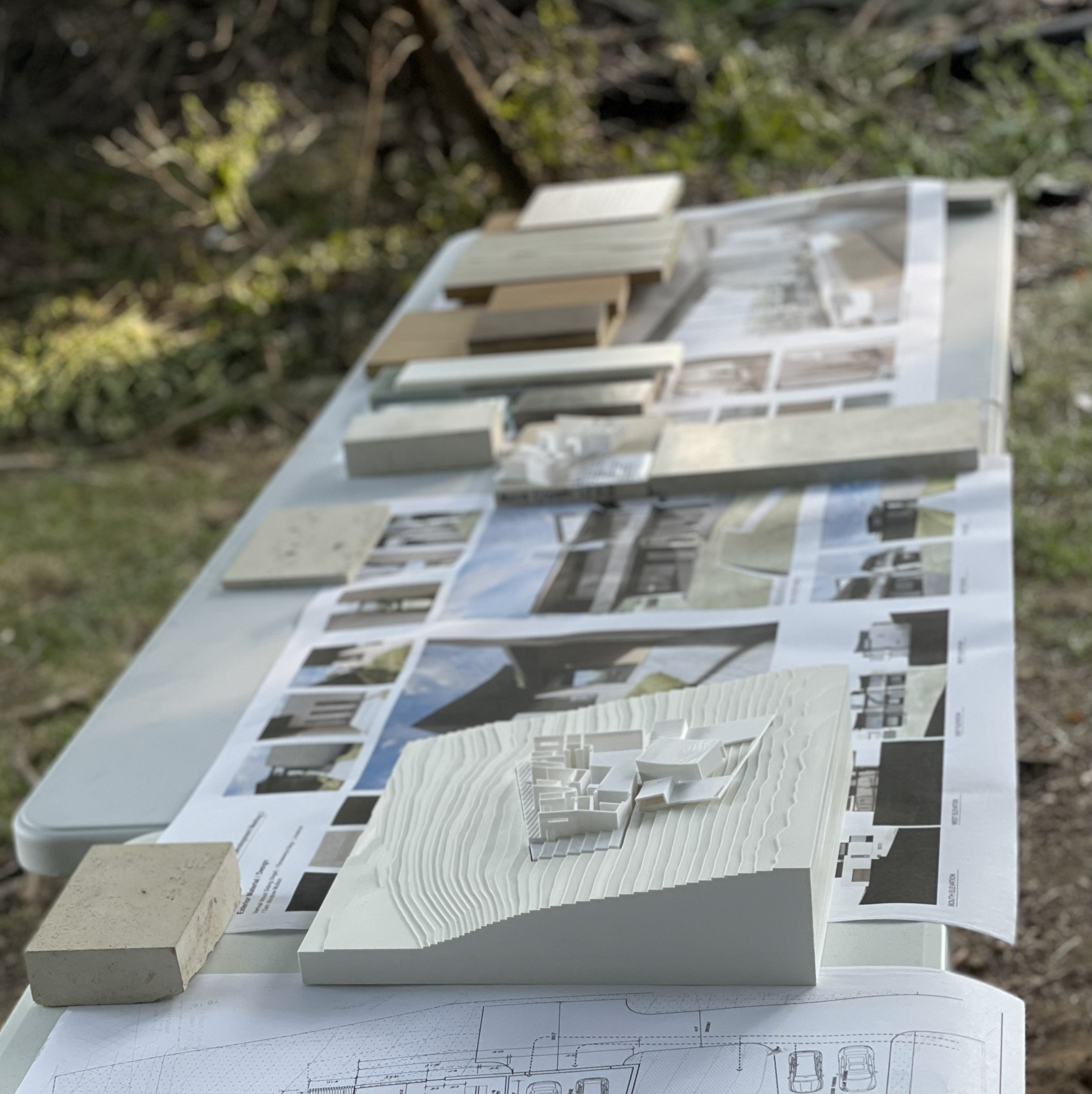 Architectural models, blueprints, and design plans laid out on a table outdoors.