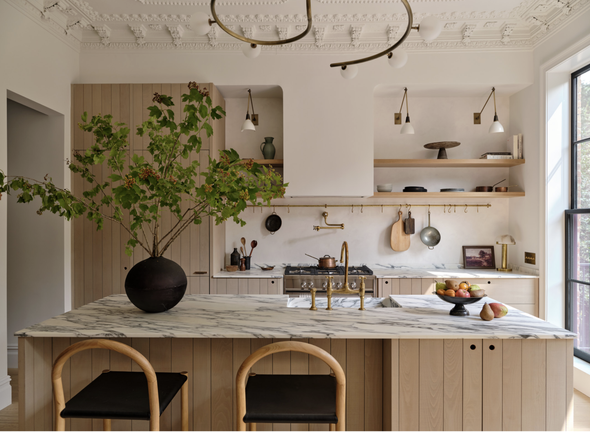 A modern kitchen with a large marble island, a black vase with leafy green branches, wooden chairs, open shelves with bowls and decorative items, a window on the right letting in natural light, a stove, and minimalistic decor.