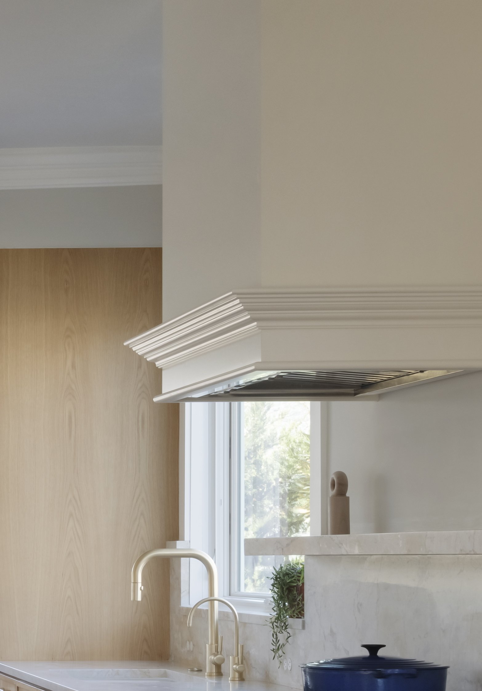 Modern kitchen with a white range hood, a marble countertop, a window with greenery outside, and a blue pot on the stove.