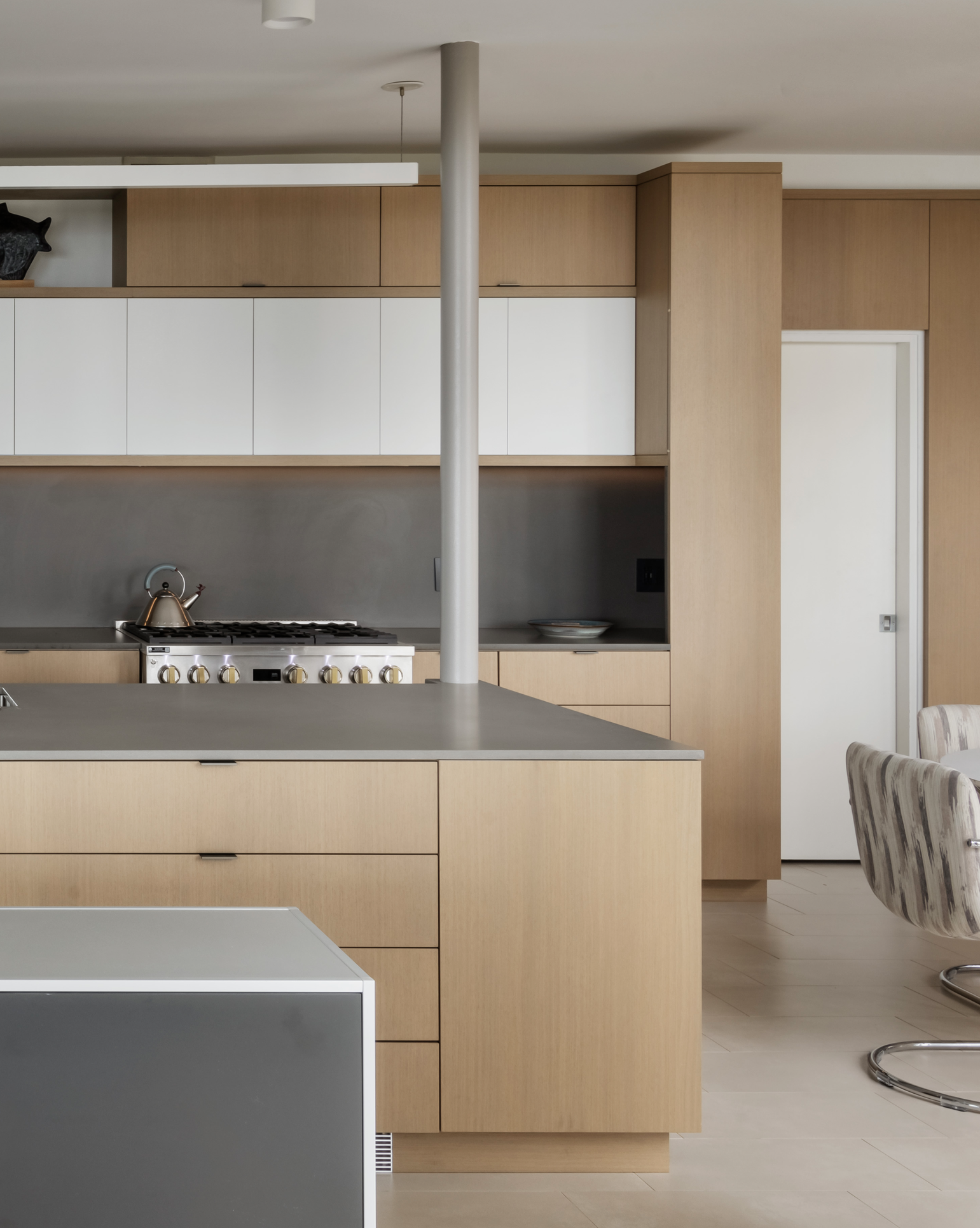 Modern kitchen with light wood cabinetry, stainless steel appliances, and a gray countertop, with a round dining chair visible on the right side.