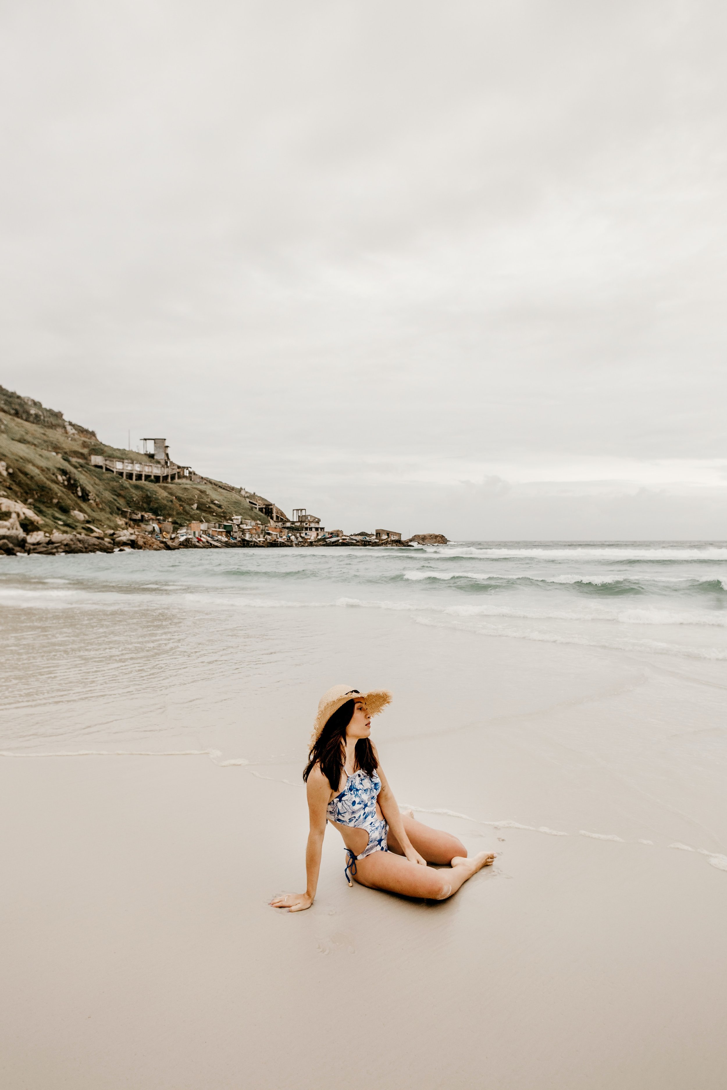 Woman in swimsuit sitting on a sandy beach with ocean waves and a cloudy sky in the background.