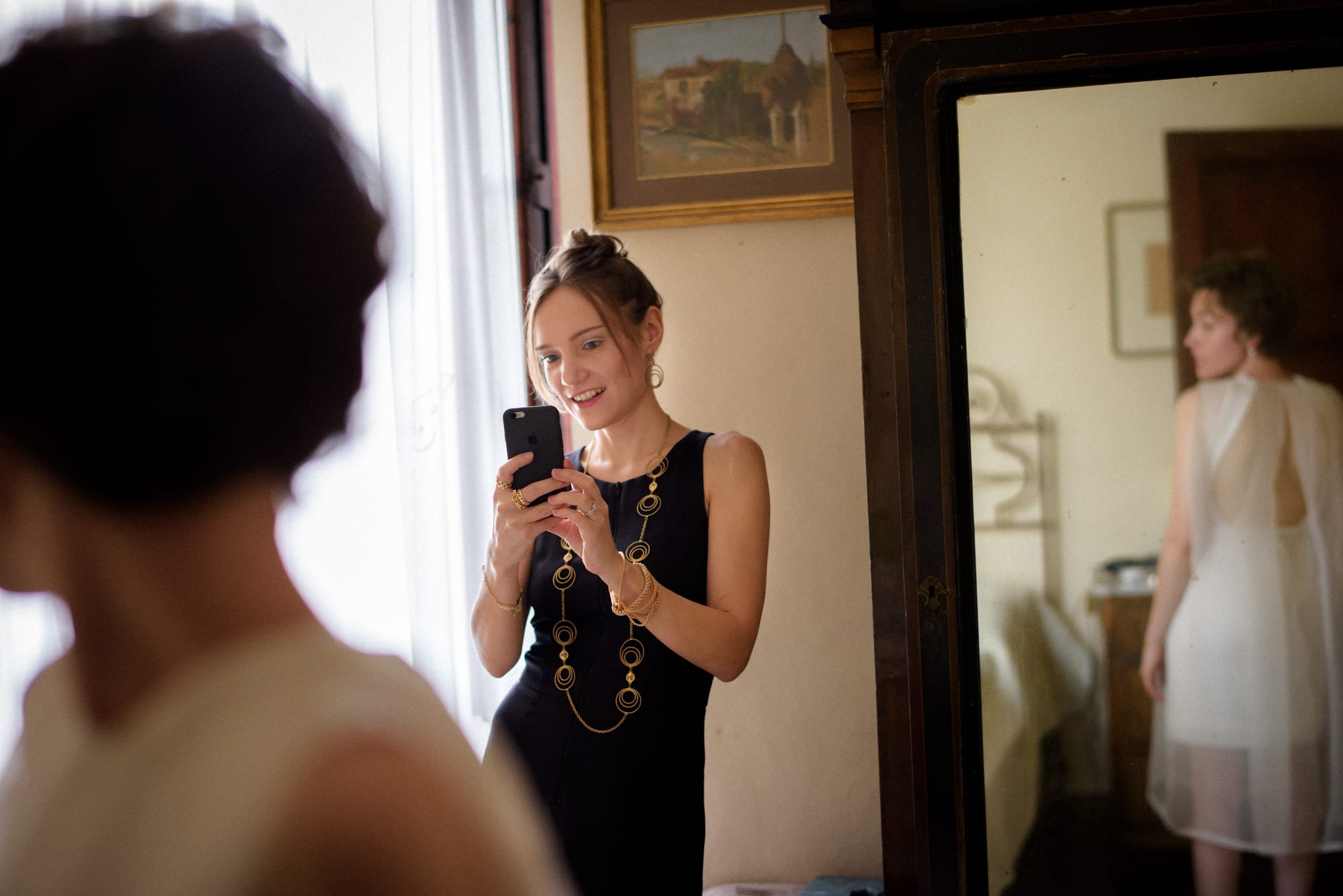 Woman in a black dress with gold jewelry taking a photo in a mirror, capturing her reflection and another person in a white dress.