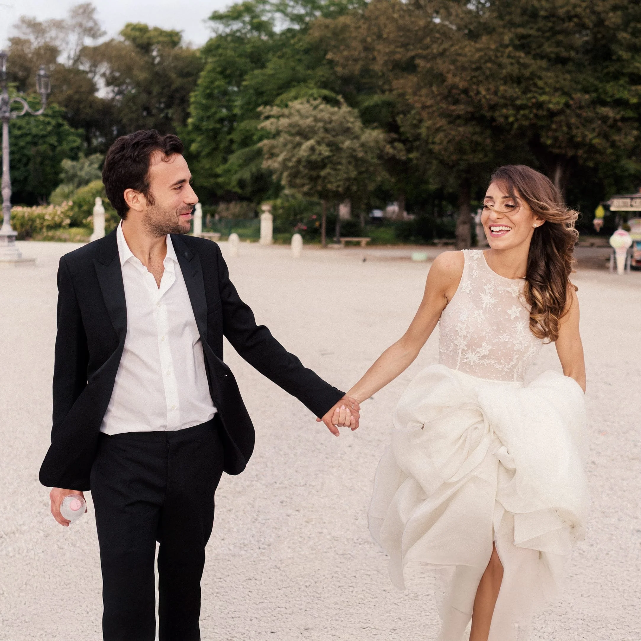 A smiling couple holding hands in formal attire; the woman is in a wedding dress and the man in a suit, walking outdoors.