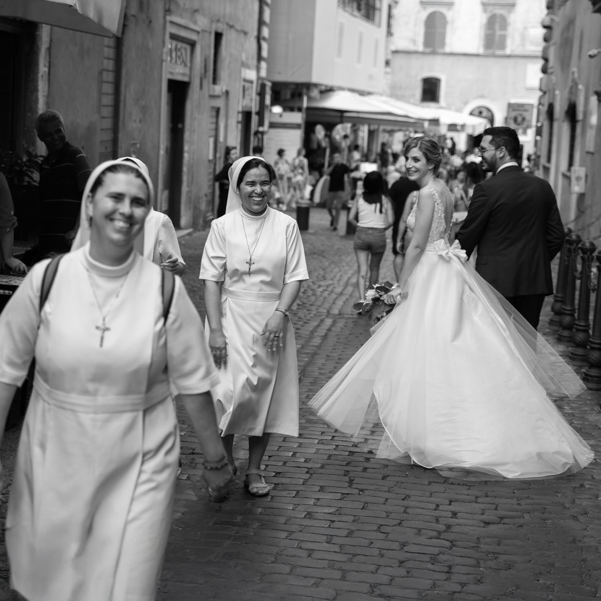 A black and white photo of a bride and groom walking on a cobblestone street, followed by nuns wearing habits. The street is lined with buildings and people in the background.