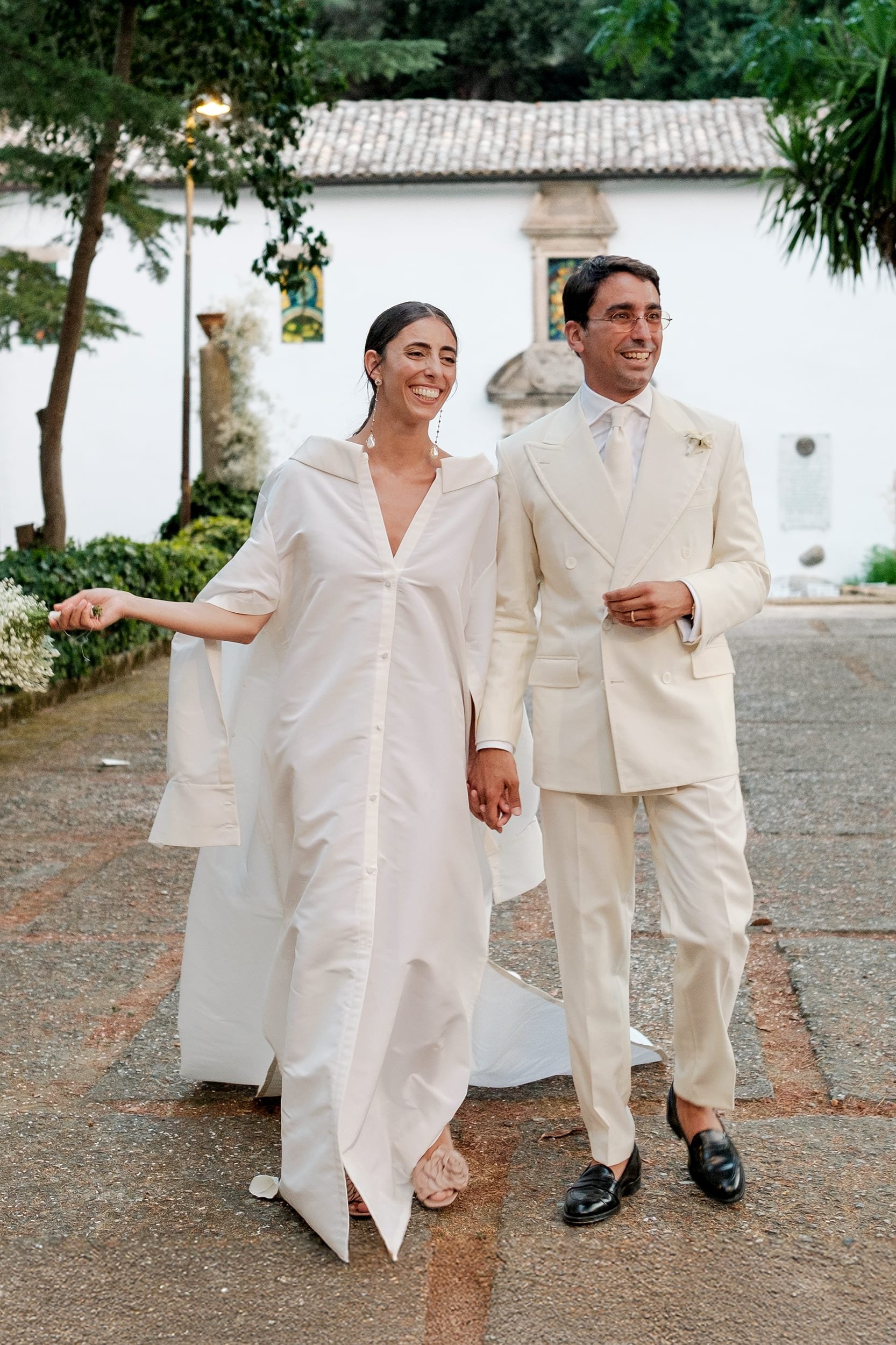 Sposi vestiti in eleganti abiti bianchi che camminano all'aperto, tenendosi per mano e sorridendo, immortalati in una scena di autentica fotografia di matrimonio.