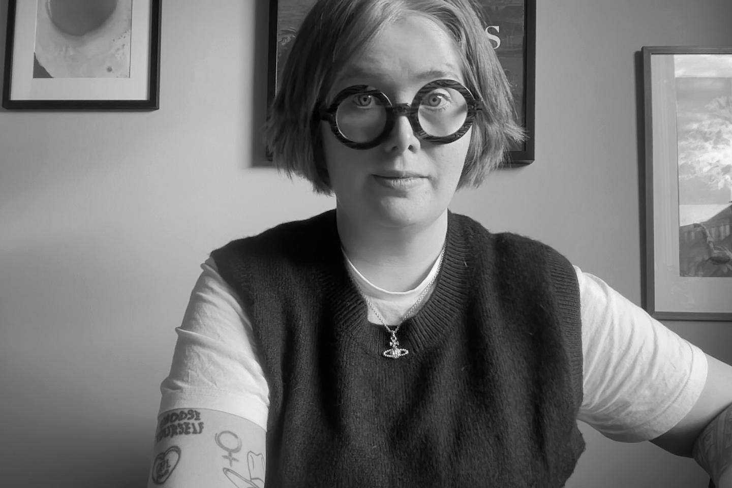 Svartvitt foto av en kvinna med kort, håret frisyr, stora runda glasögon, vita t-shirt och svart väst, sittande vid ett bord i ett rum med konst på väggarna.