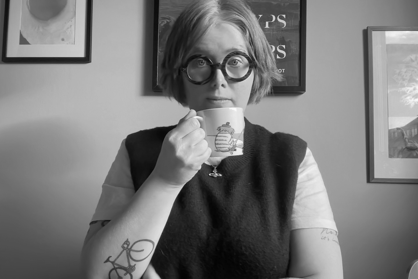 En person med kort hår och stora runda glasögon som håller en mugg med en tecknad mus på. De är inomhus, vita väggar med några målningar eller fotografier i ramar i bakgrunden.