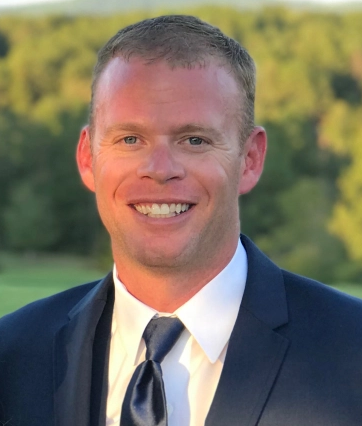 Man in a suit smiling outdoors against a blurred natural background