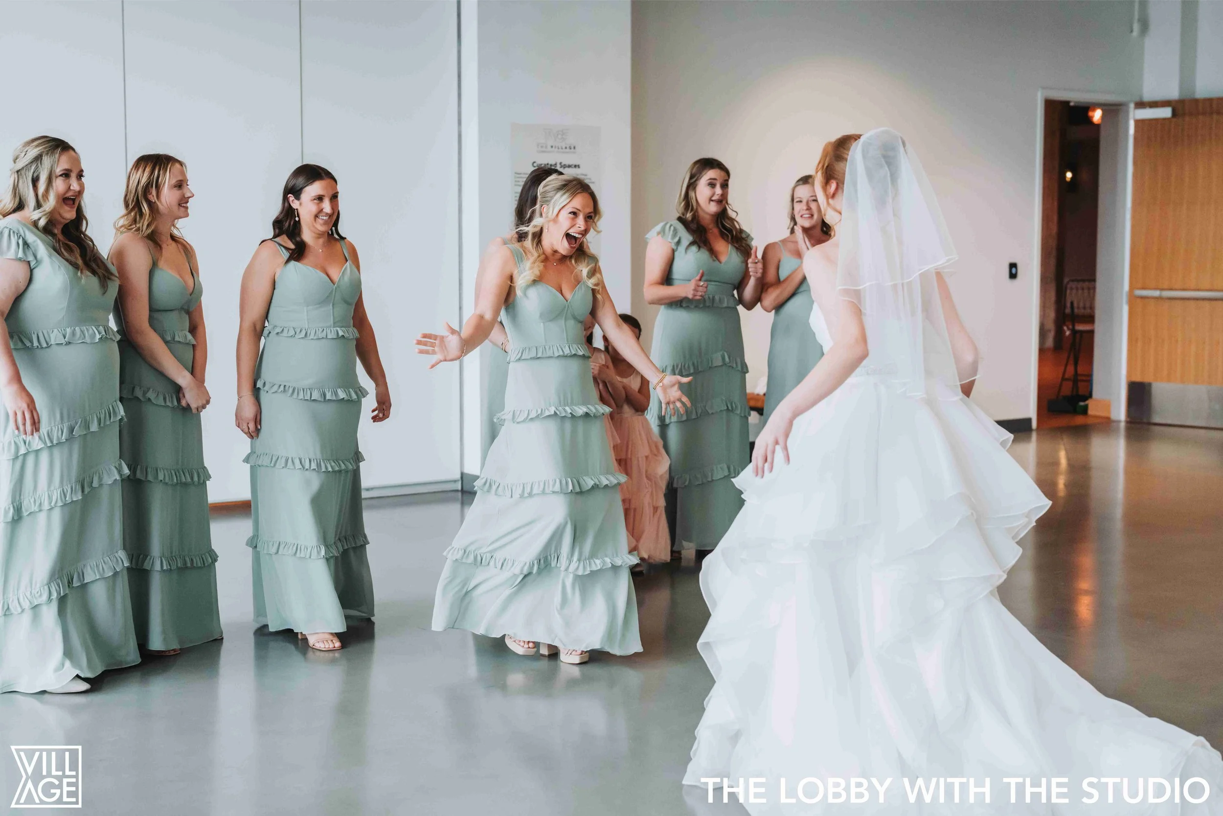 Bride in white wedding dress and veil dancing in front of a group of smiling bridesmaids in matching sage green dresses in a bright indoor venue.