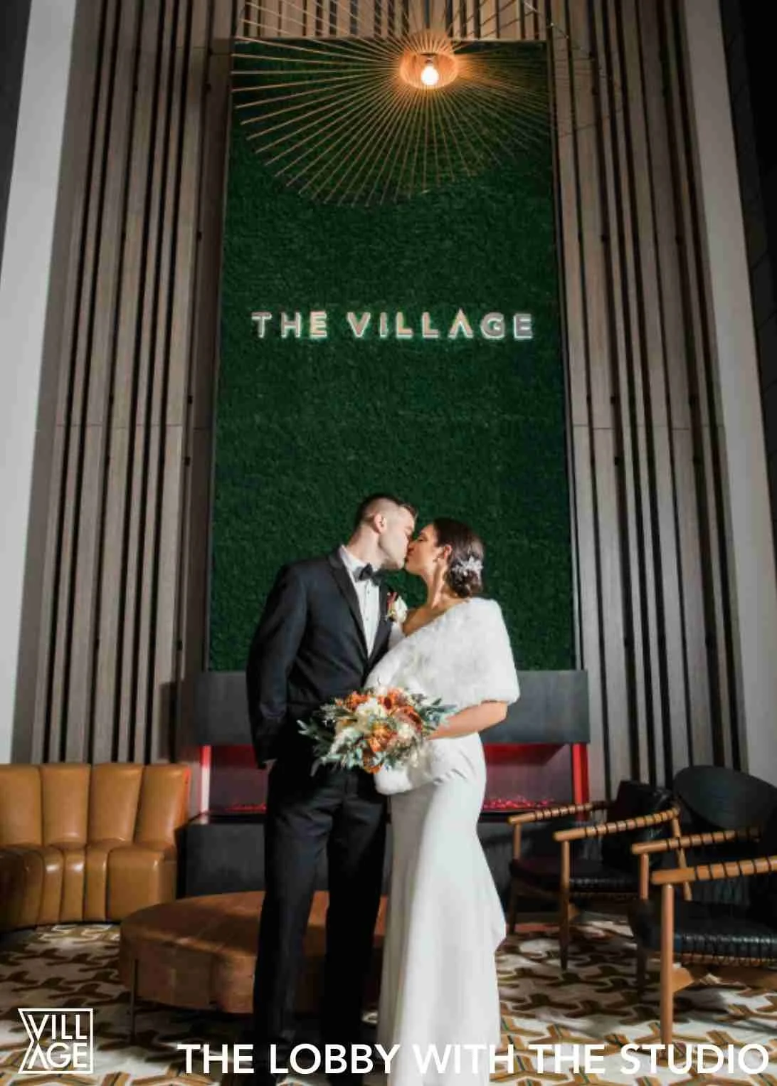 A bride and groom sharing a kiss in a modern lobby with green wall and decorative lighting, with the sign 'The Village' above them.