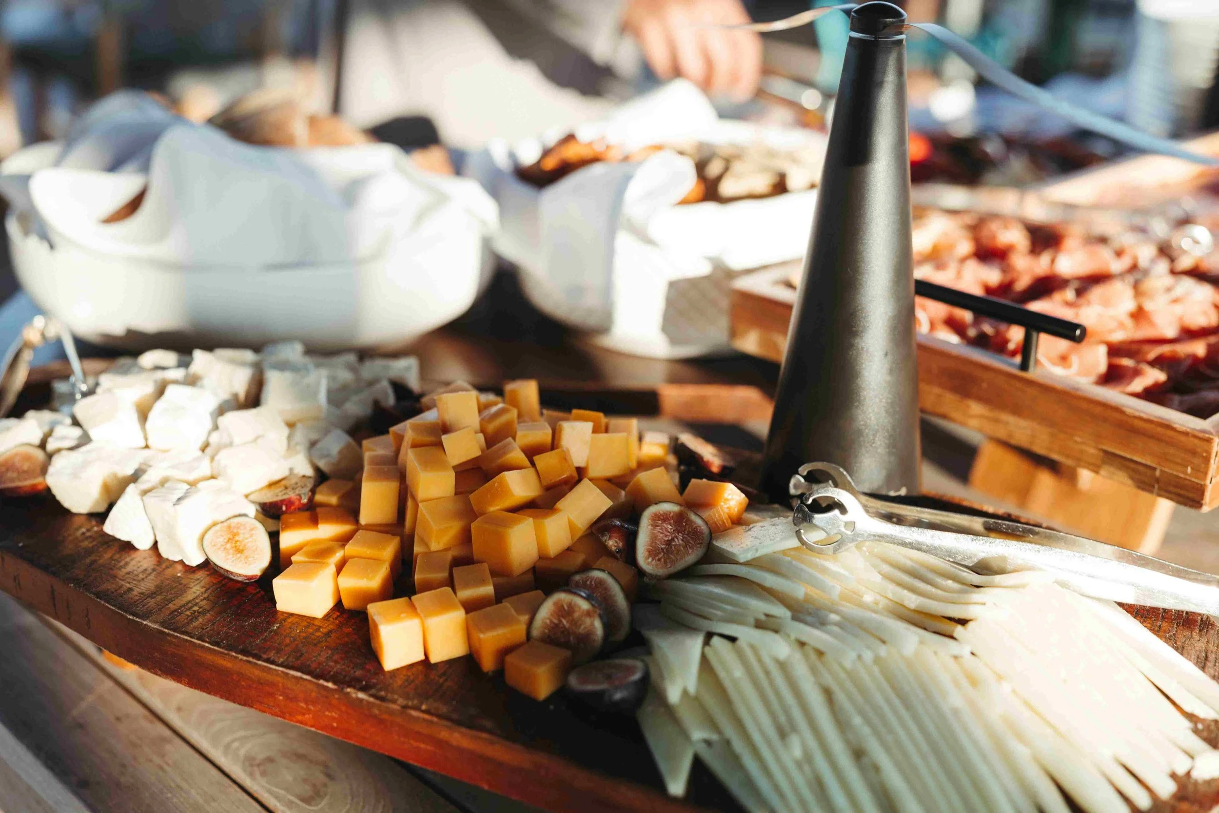 Assorted cheeses, sliced and cubed, arranged on a wooden table at a food event, with a black cone-shaped object and a pair of cheese tongs.