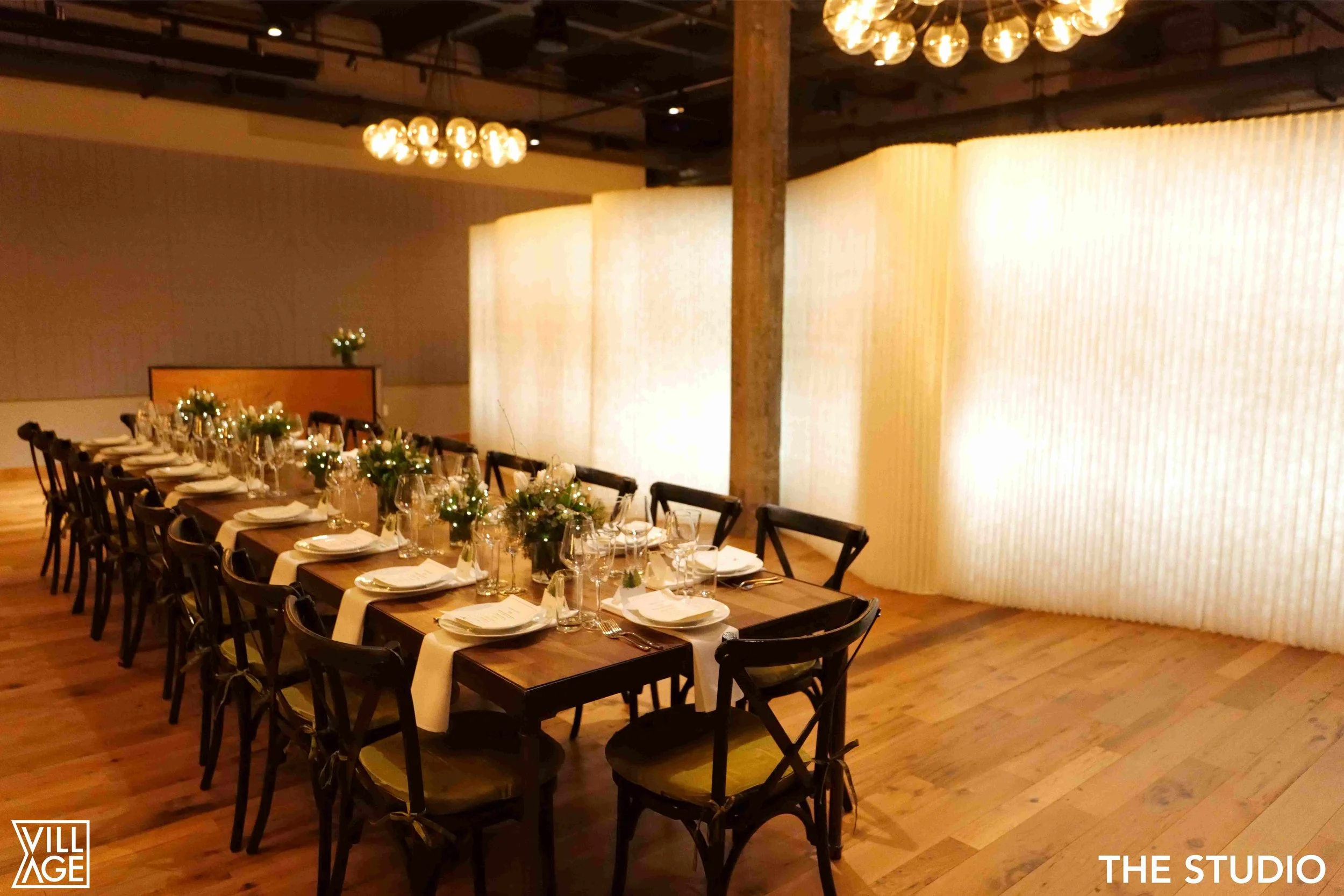 Long wooden table set for a formal dinner with white napkins, multiple wine glasses, plates, and silverware, decorated with green leafy centerpieces and white flowers, in a warmly lit event space with a large illuminated white backdrop and ceiling li