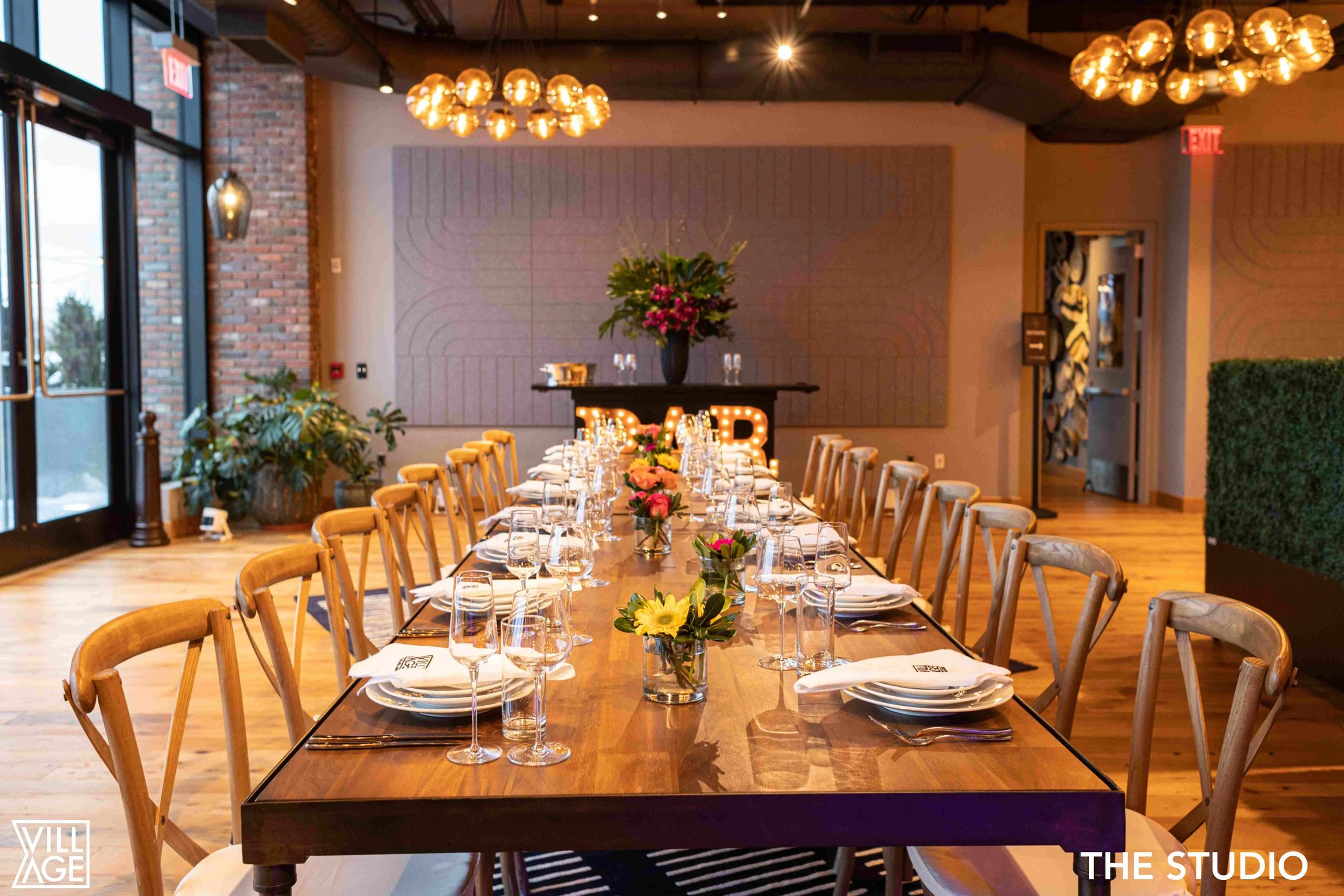 A long wooden dining table set for a formal event with wine glasses, plates, and floral centerpieces inside a modern, stylish restaurant or event space with wooden floors, large windows, and warm lighting fixtures.