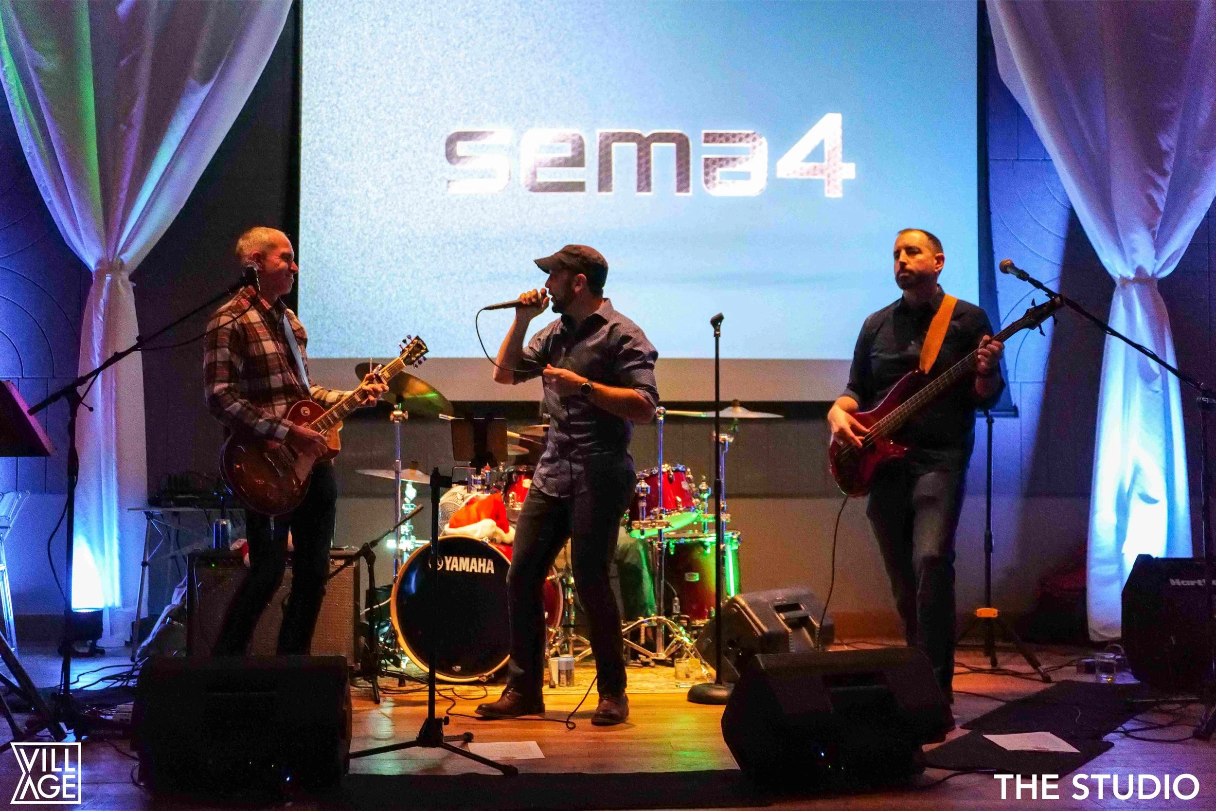 A band performing on stage with a bass guitar, electric guitar, and drums, with a large screen behind showing 'Sema4'.