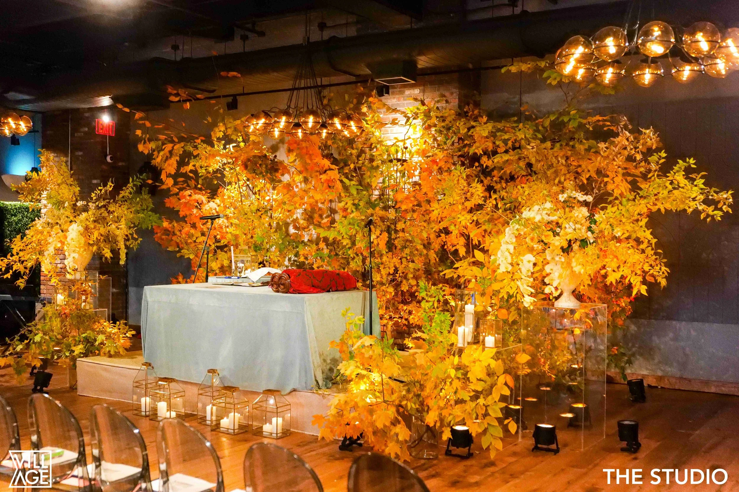 A decorated event space with fall-themed decorations, large floral arrangements, candles in glass lanterns, and a DJ or music setup with turntables and a speaker, under warm lighting fixtures.