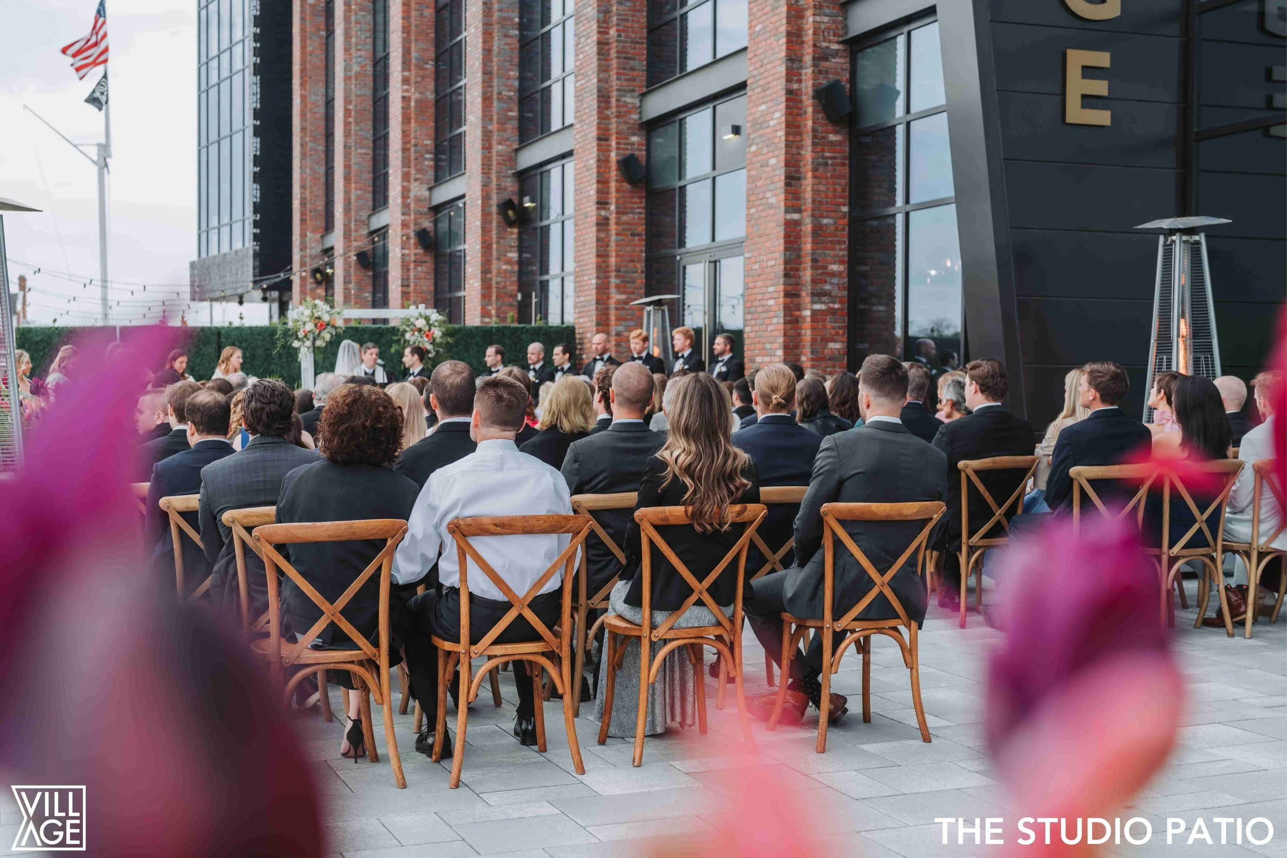 Outdoor wedding ceremony on a patio with guests seated in wooden chairs facing a couple and officiant at an altar decorated with flowers, set against a brick building.