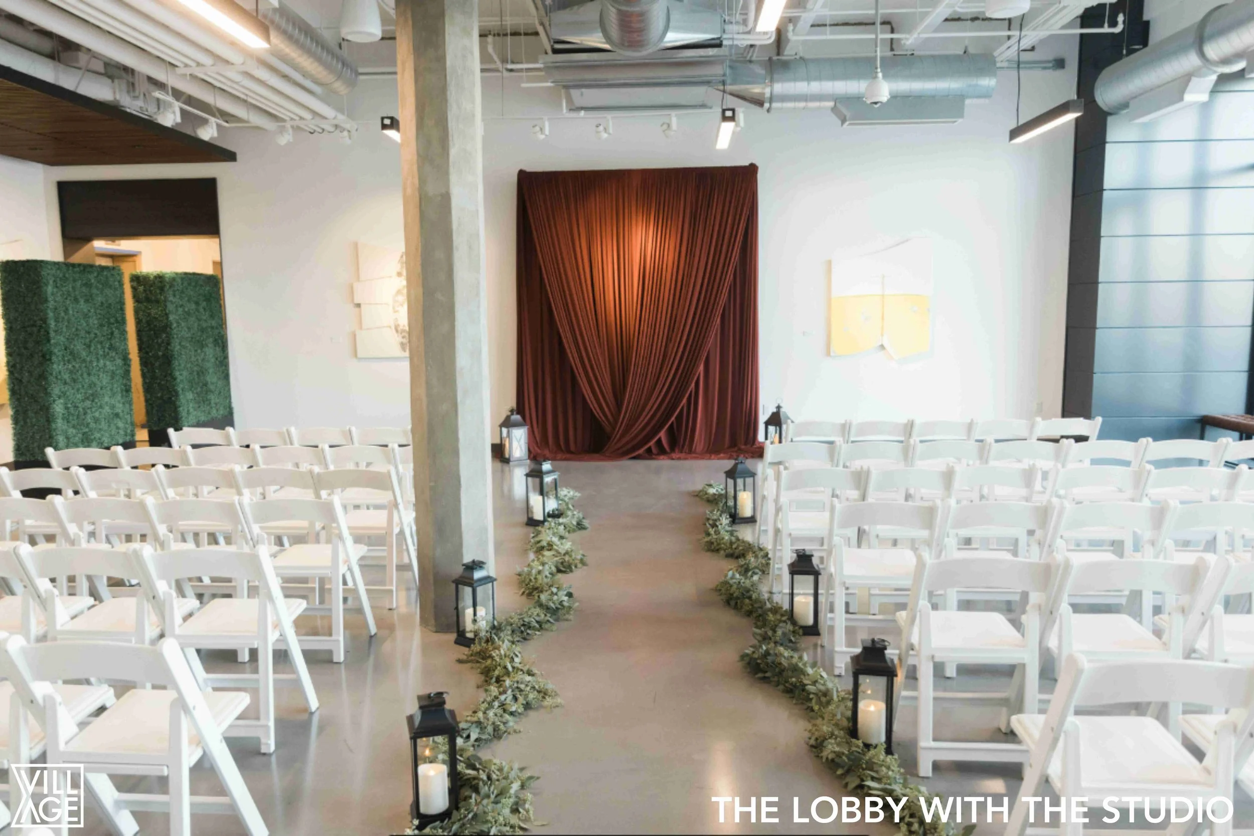 Indoor wedding ceremony setup with rows of white chairs. A central aisle decorated with candles and greenery leads to a red curtain backdrop.