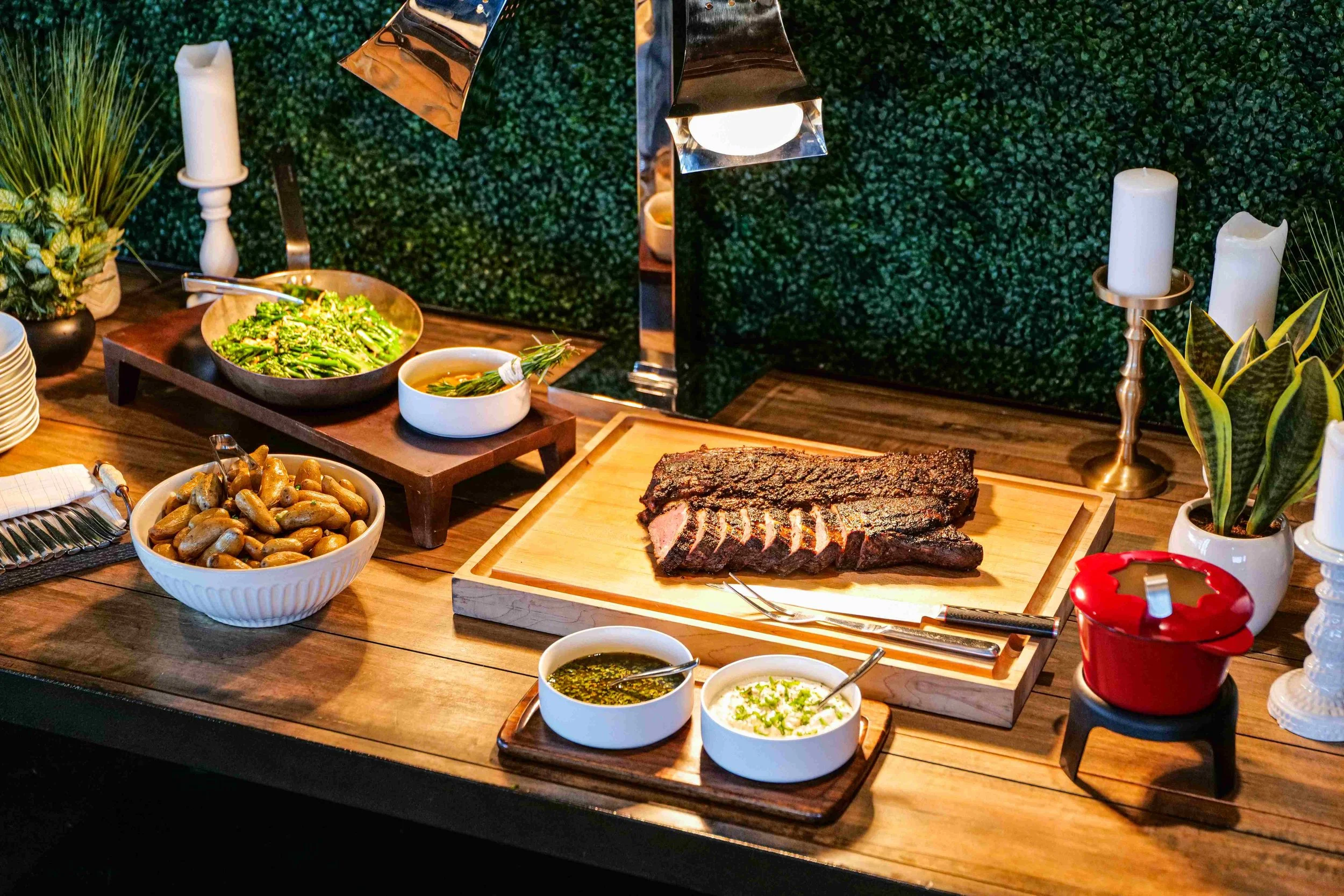 A wooden table with a large cut of cooked steak on a wooden cutting board, surrounded by bowls of beans, sauces, a salad, candles, and potted plants.
