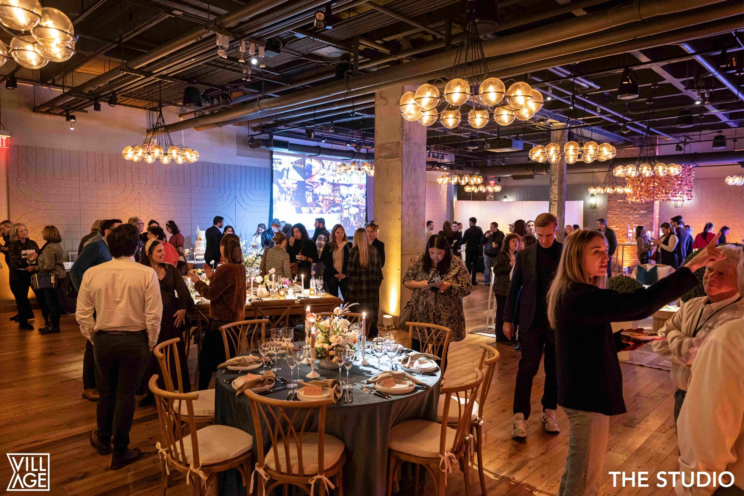Indoor event with people gathering around decorated tables, warm lighting, and a large digital screen on the back wall of a venue.
