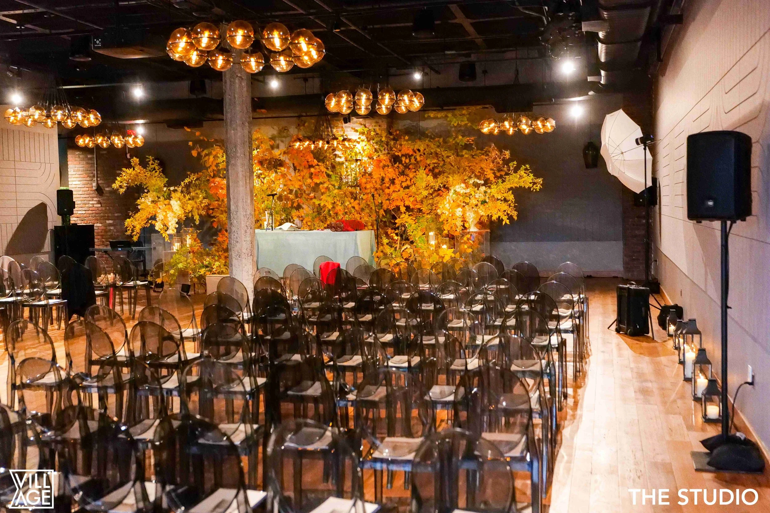 Indoor event space with rows of transparent chairs facing a stage decorated with large orange and yellow autumn leaves and candles, with warm overhead lighting.