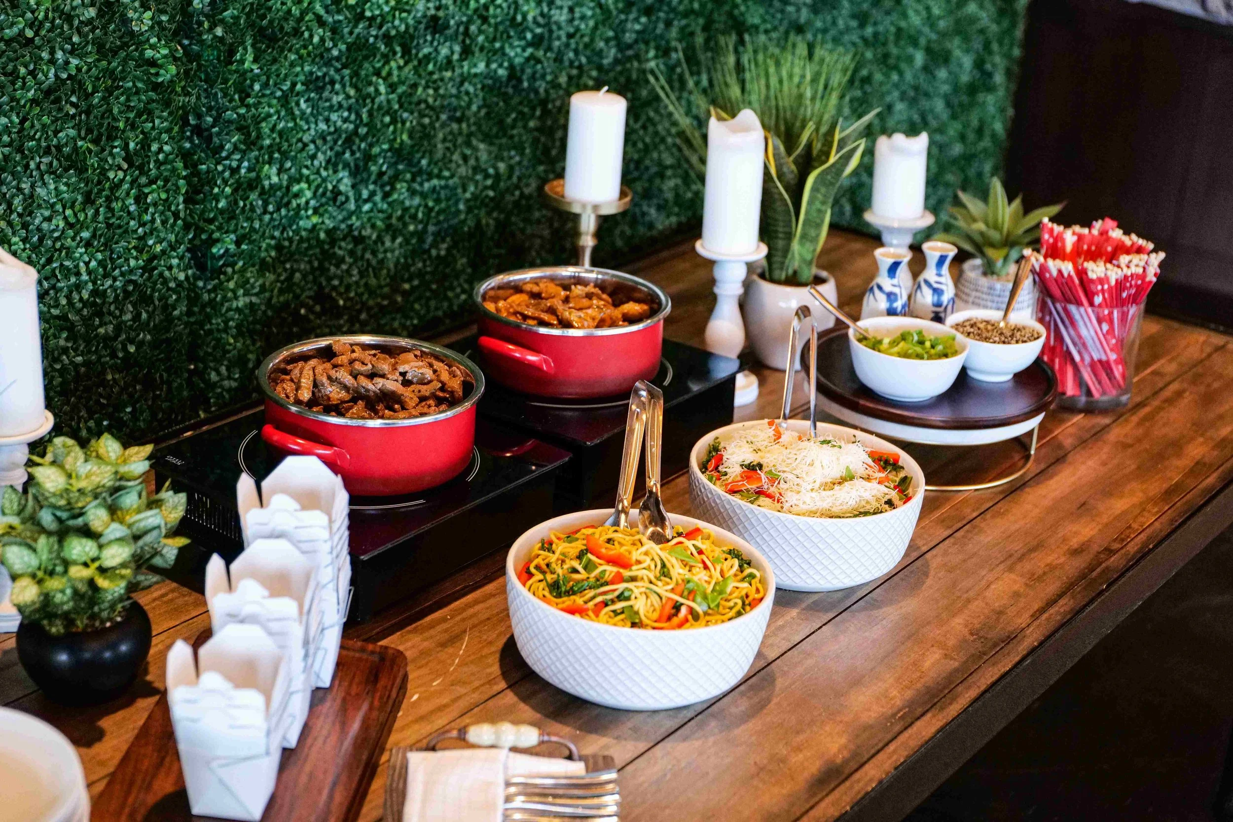 Buffet spread with two pots of cooked meat, two large bowls of noodles and salad, small bowls of green onions and seeds, and a container of red straws on a wooden table decorated with candles and small plants.