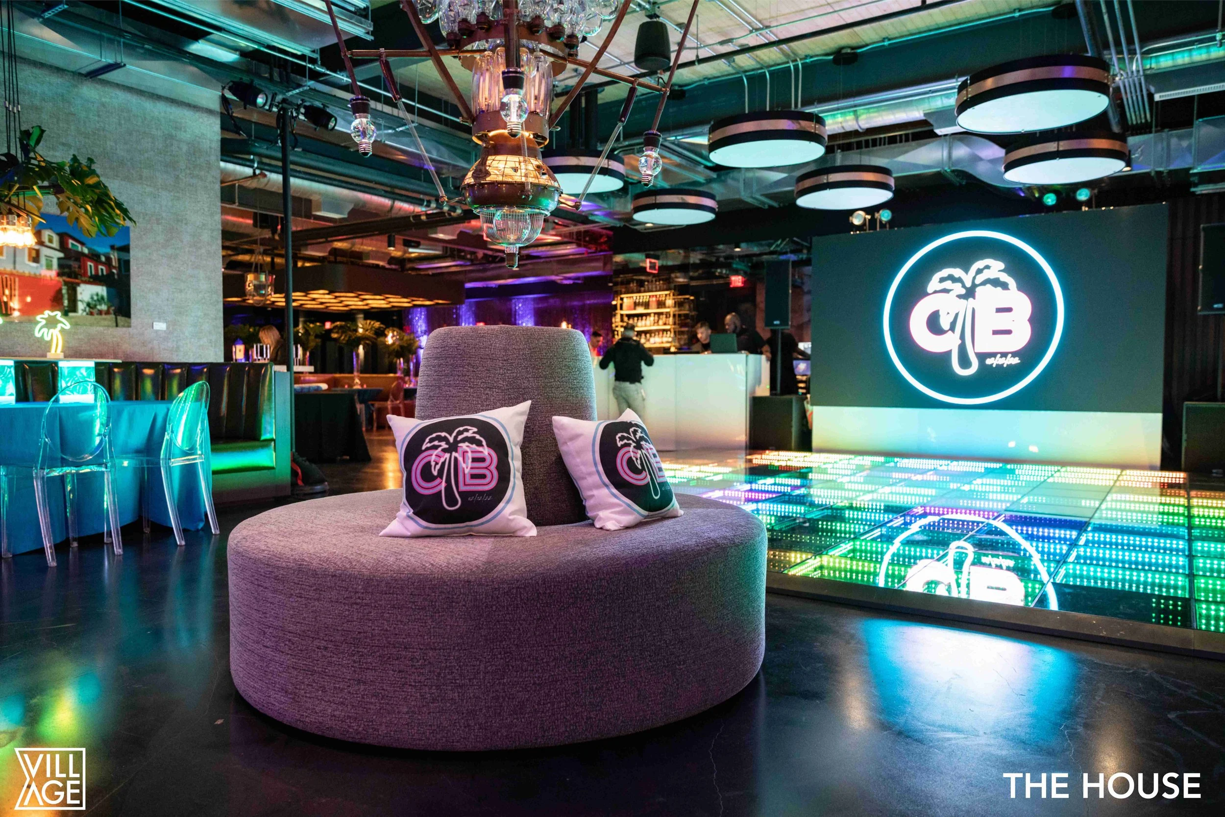 Interior of a modern nightclub with colorful lighting, a lounge chair with palm tree pillows, a dance floor with LED lights, and a neon sign displaying the nightclub's logo, 'C B' with a palm tree.