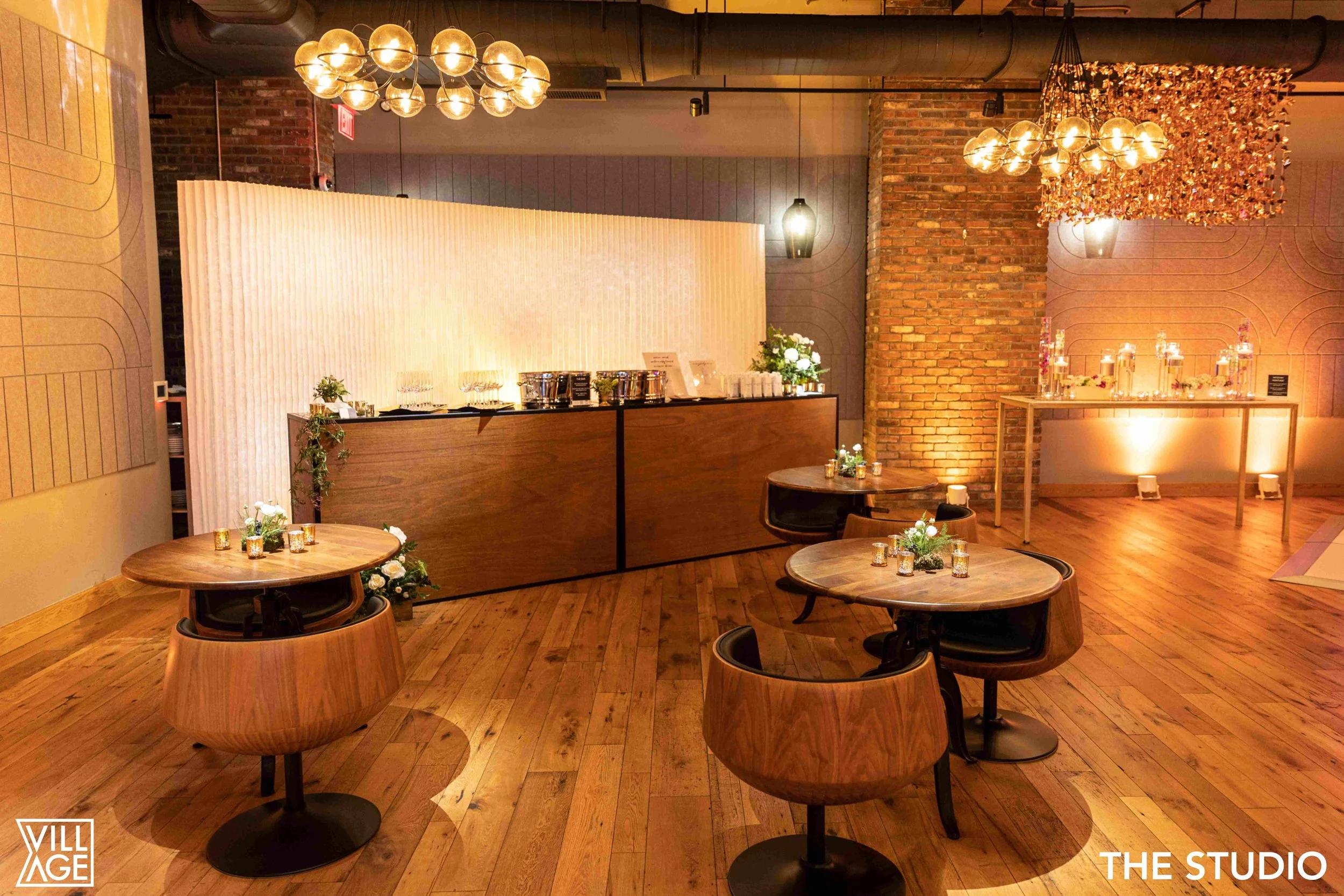 Elegant event space with wooden floors, round tables with small floral arrangements, a bar area with drapes and drinks, and modern chandeliers with warm lighting.