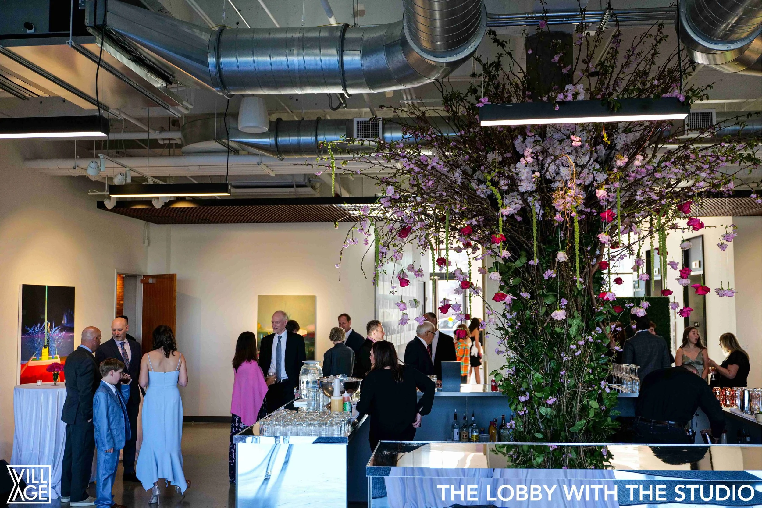 A modern art gallery lobby with a large floral installation and people chatting and mingling. Artwork hangs on the walls, and a bar area is visible. The bottom right corner has text that reads 'THE LOBBY WITH THE STUDIO' and the bottom left corner ha