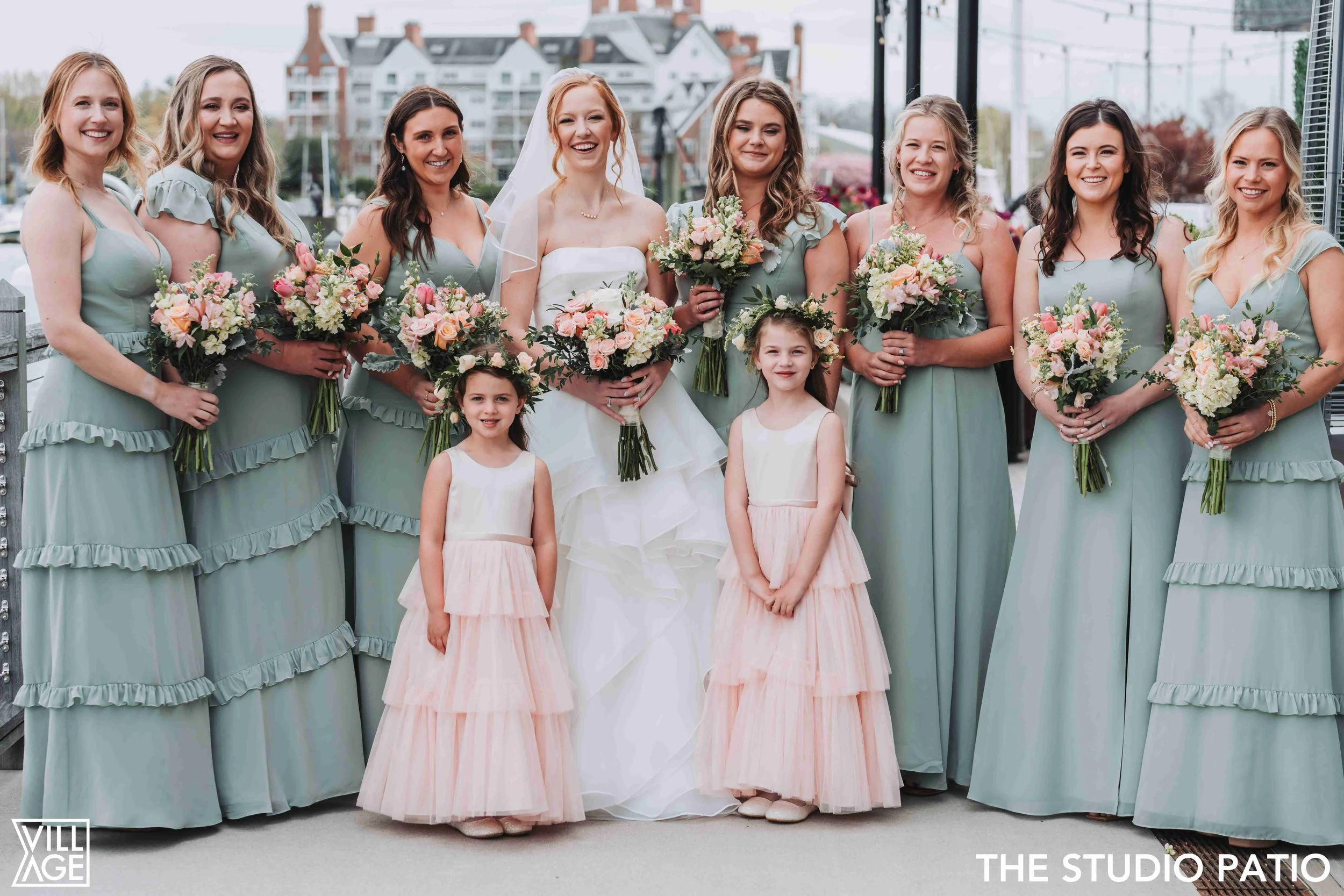 Group of women in matching green dresses, celebrating a wedding, with two young girls in pink dresses, all holding bouquets of flowers, standing outdoors on a balcony or patio.