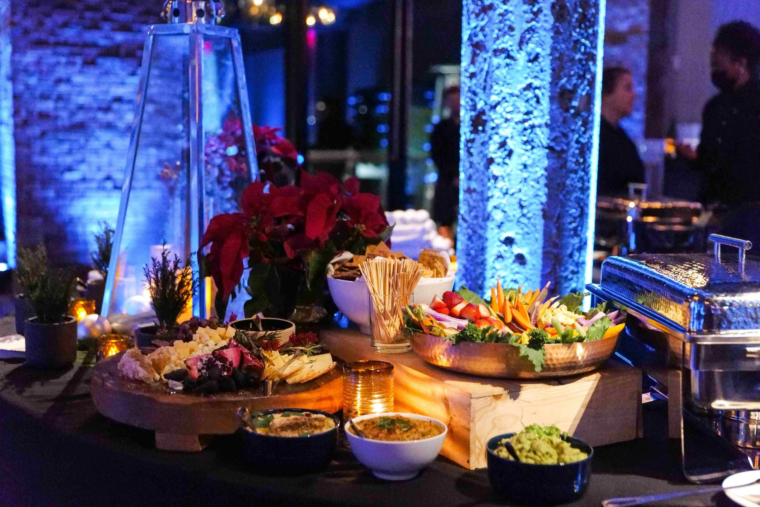 Buffet table with vegetables, cheese, crackers, and dip, decorated with red poinsettias and small potted plants, at a festive event with blue lighting and in the background people socializing.