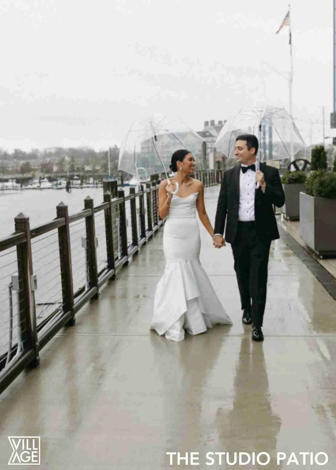 Bride and groom walking hand in hand on a rainy day, each holding a clear umbrella, on a patio overlooking a body of water, with an American flag in the background.