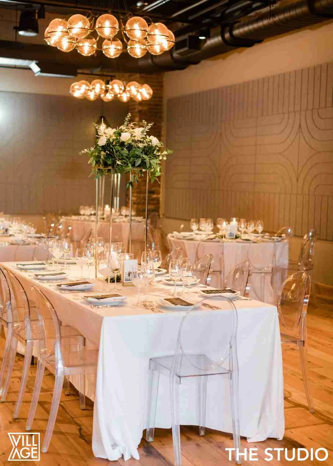 Elegant wedding reception setup with a long table, clear acrylic chairs, white tablecloth, wine glasses, and a tall floral centerpiece featuring white roses and greenery. Warm lighting from modern globe chandeliers hangs above.