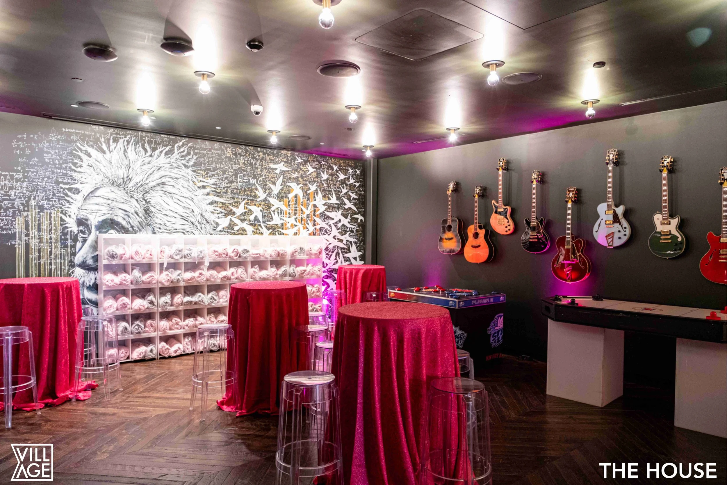 An indoor space with red velvet tables and clear chairs, black wall with hanging guitars, a wall with a lion mural and bird illustrations, and a foosball table.