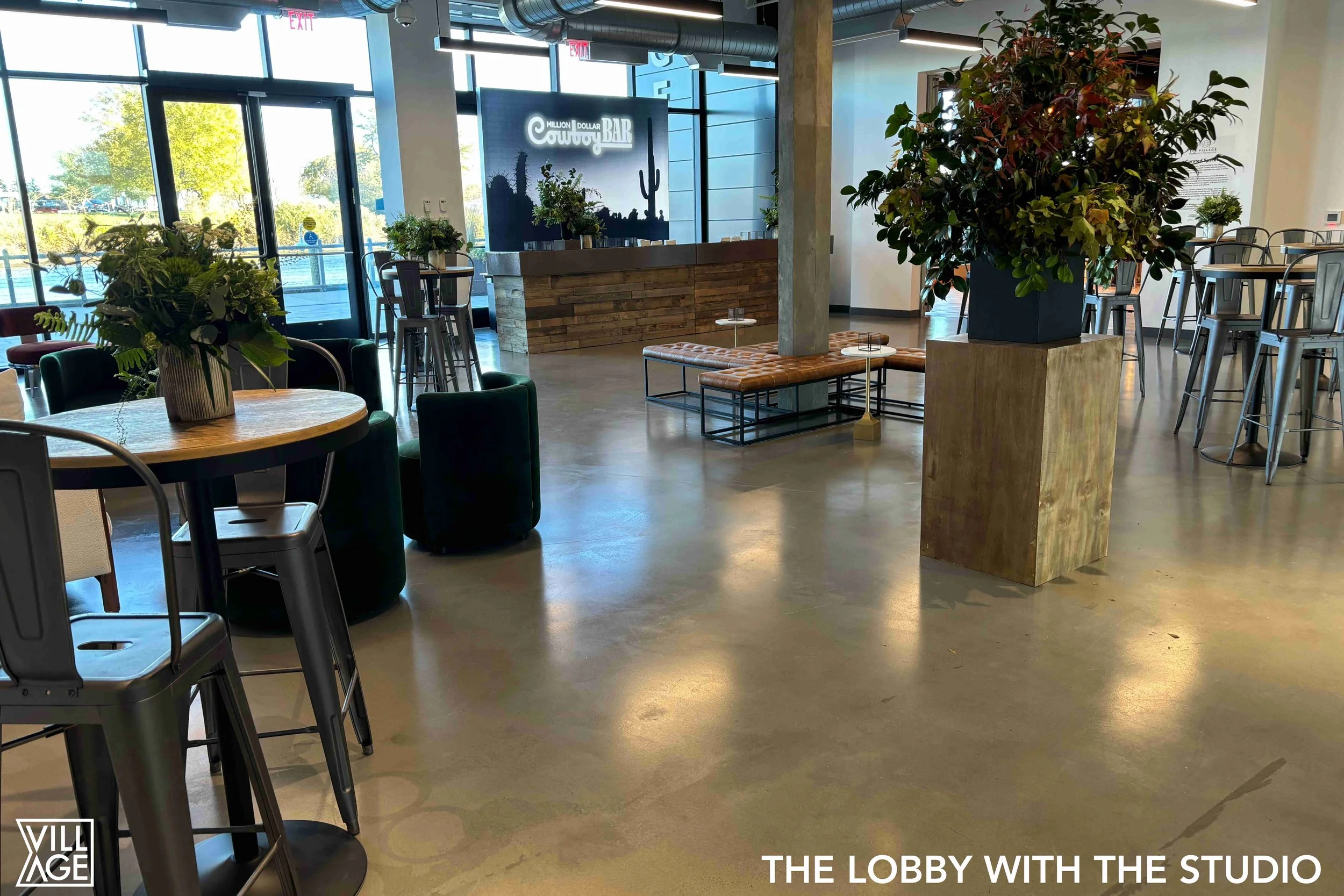 Modern lobby with large plants, a wooden counter with a digital screen, and seating areas including round tables with chairs and a tufted leather bench by a column. Large windows let in natural light, and there are high stools near a bar-height table