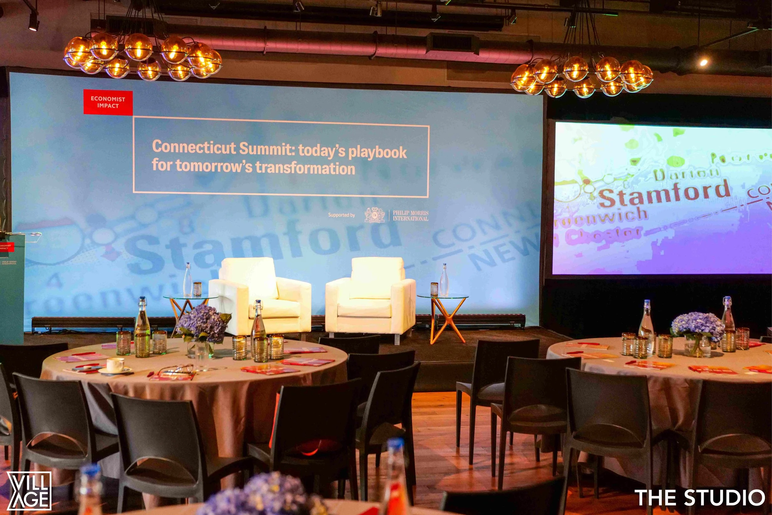 Event space with tables set for a conference, with round tables covered with pink tablecloths, water bottles, glasses, and floral centerpieces. There is a stage in the background with two white chairs, a small orange side table, and a large screen di