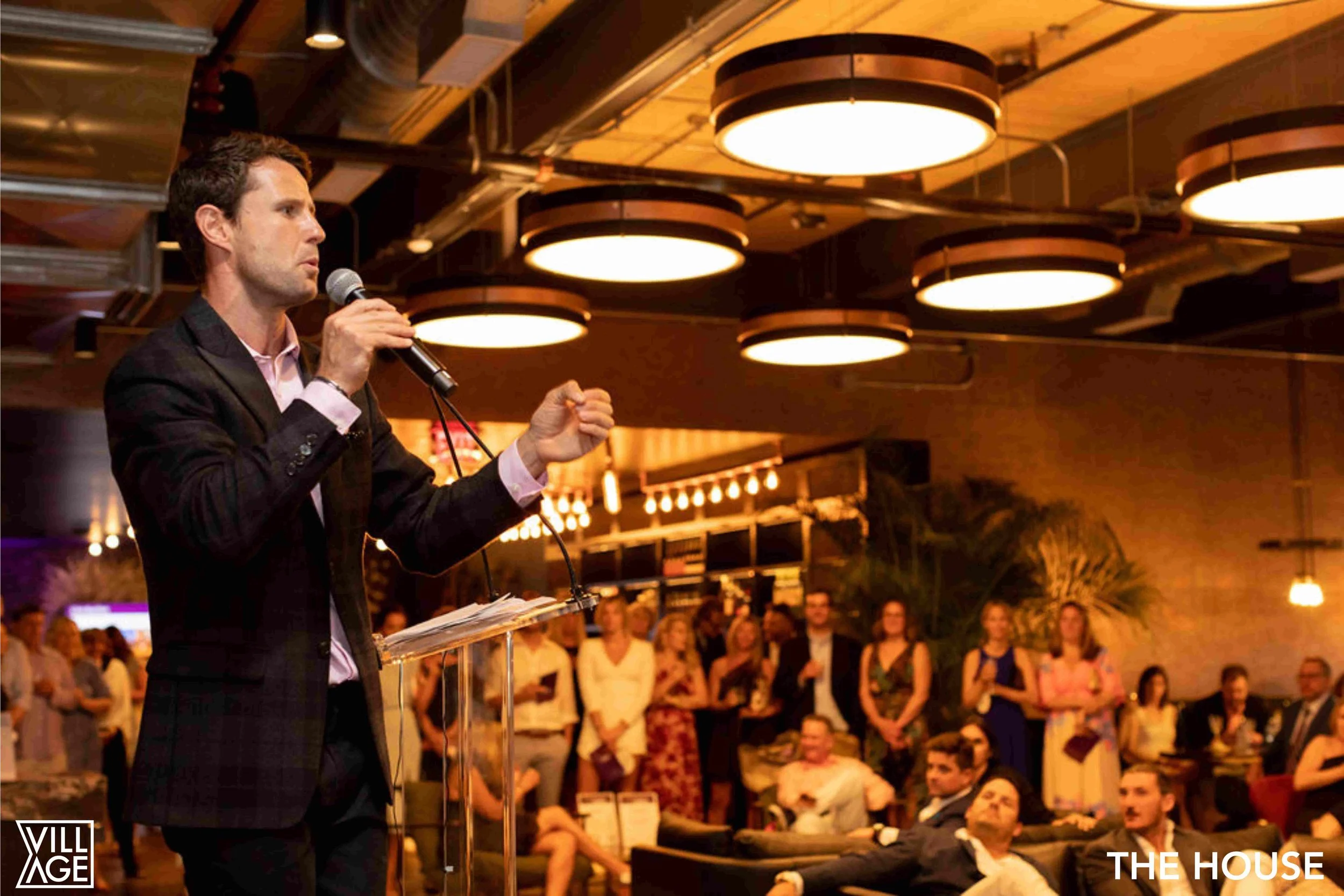A man in a suit speaking into a microphone at an indoor event with an audience seated and standing behind him, in a warmly lit room with modern circular ceiling lights.