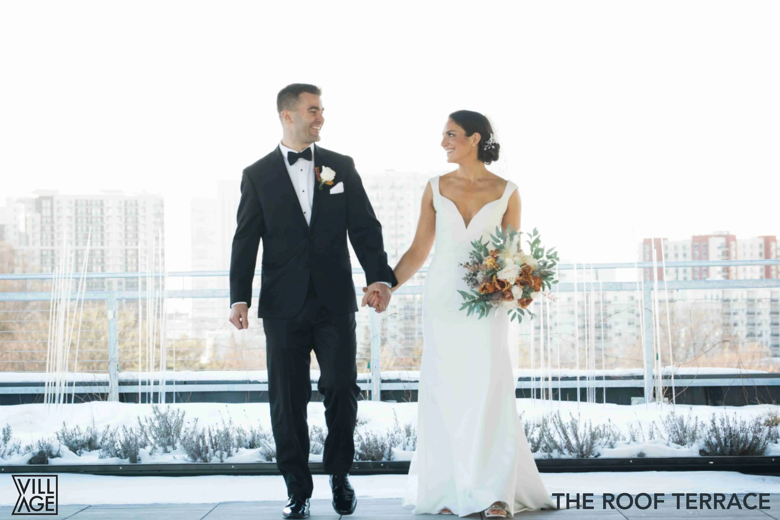 A bride and groom walking hand-in-hand on an outdoor terrace with snow and cityscape in the background, smiling at each other after their wedding.