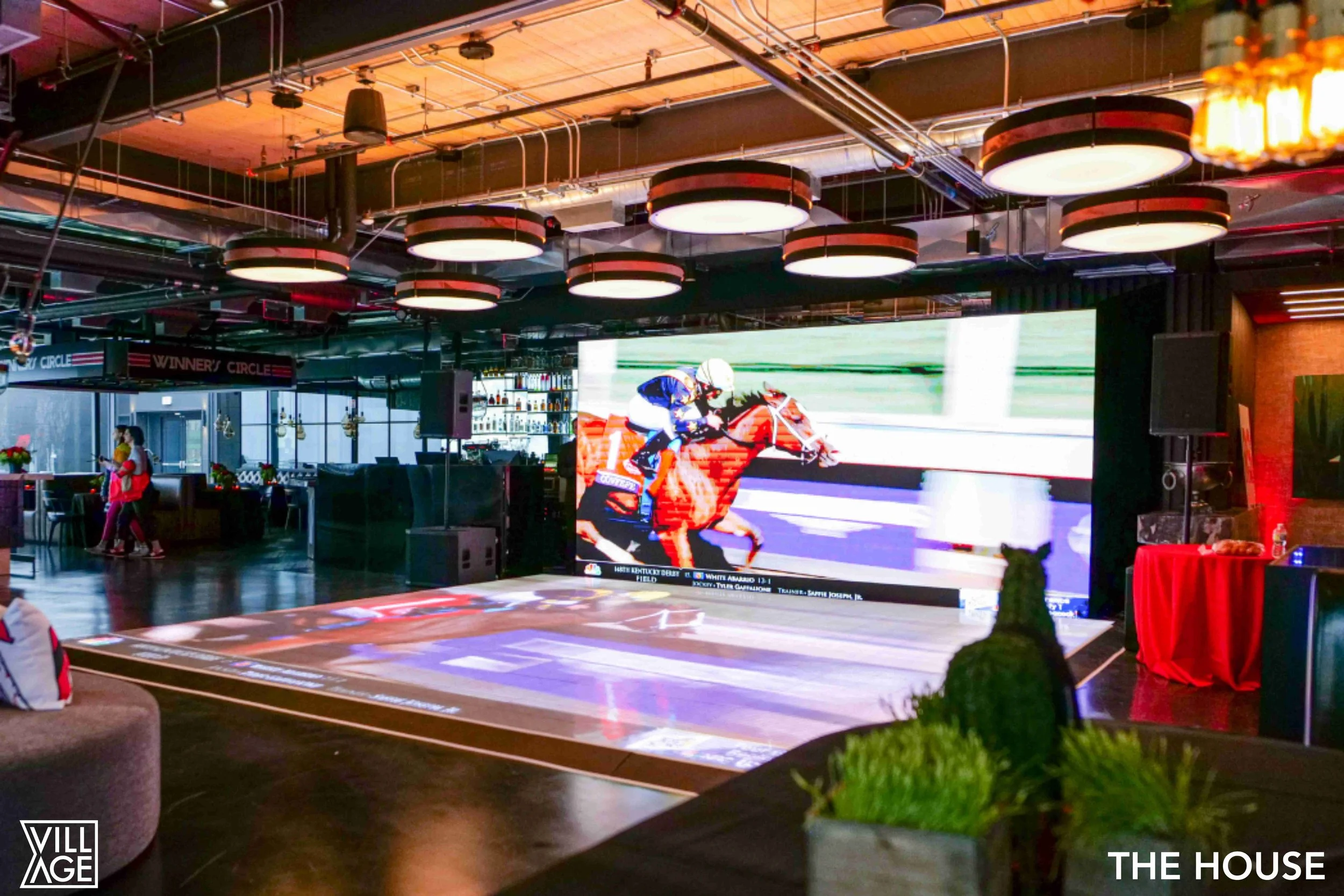 Indoor event space with a large screen showing a horse race, a section labeled 'WINNER'S CIRCLE', some people walking, and modern lighting fixtures.